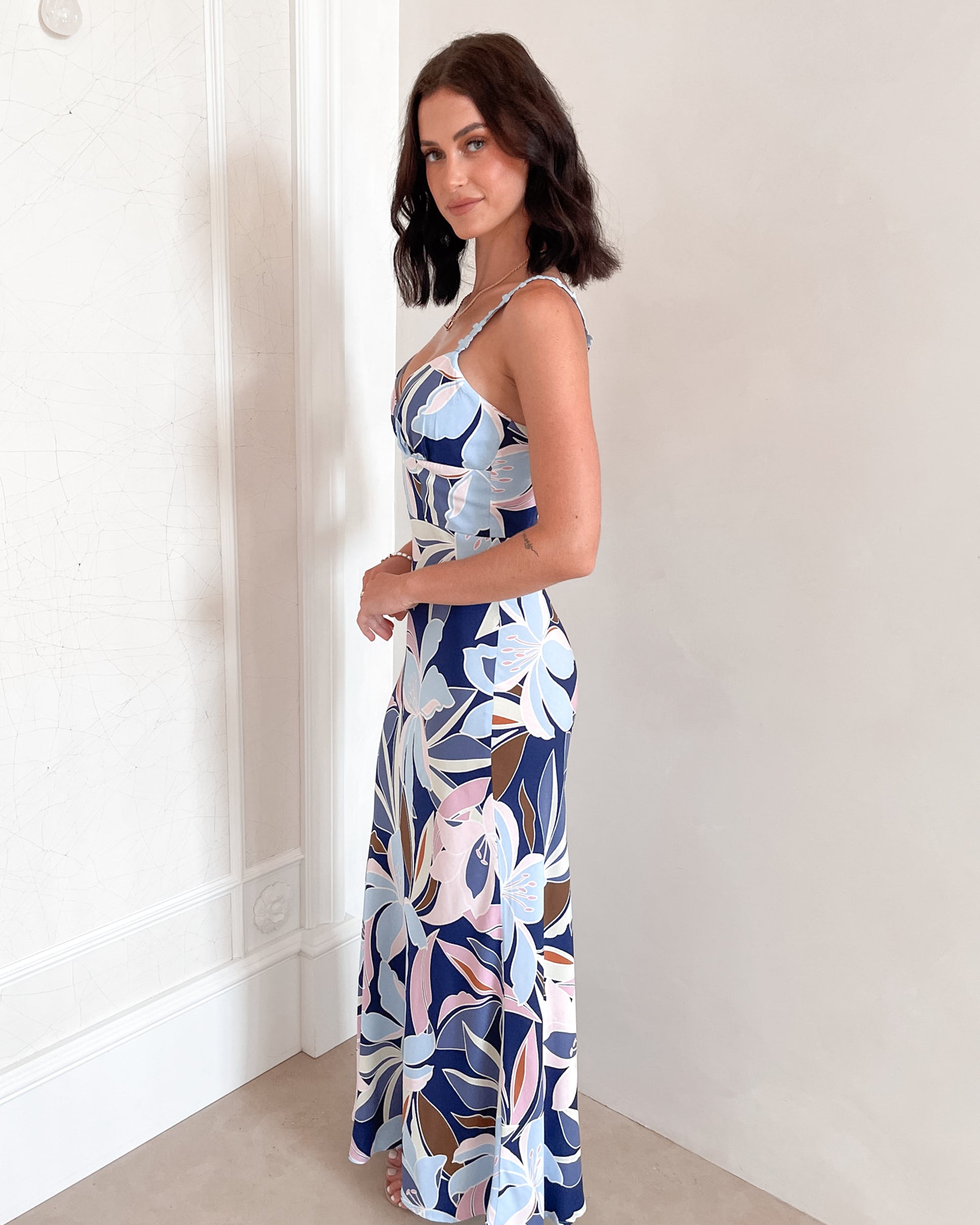 A woman stands smiling indoors against a light-colored wall, wearing a fitted, floor-length dress with a bold floral print in shades of navy blue, light blue, pink, and white. The dress features a deep V-neckline and light blue floral straps. Her dark, wavy hair is styled down, and she accessorizes with a simple gold necklace and rings. She poses confidently with her hands on her hips and is barefoot, adding to the relaxed and elegant vibe of the scene