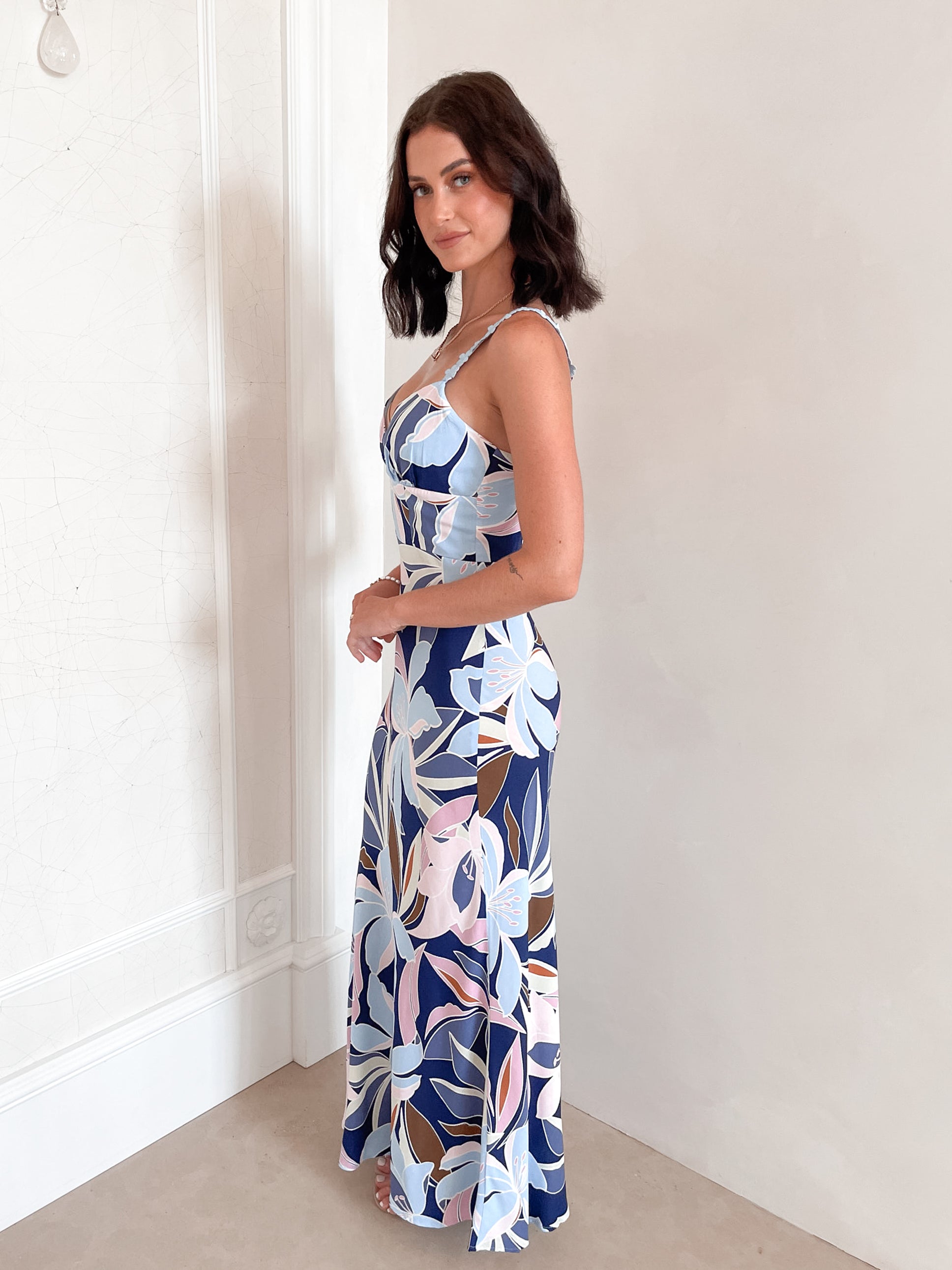 A woman stands smiling indoors against a light-colored wall, wearing a fitted, floor-length dress with a bold floral print in shades of navy blue, light blue, pink, and white. The dress features a deep V-neckline and light blue floral straps. Her dark, wavy hair is styled down, and she accessorizes with a simple gold necklace and rings. She poses confidently with her hands on her hips and is barefoot, adding to the relaxed and elegant vibe of the scene