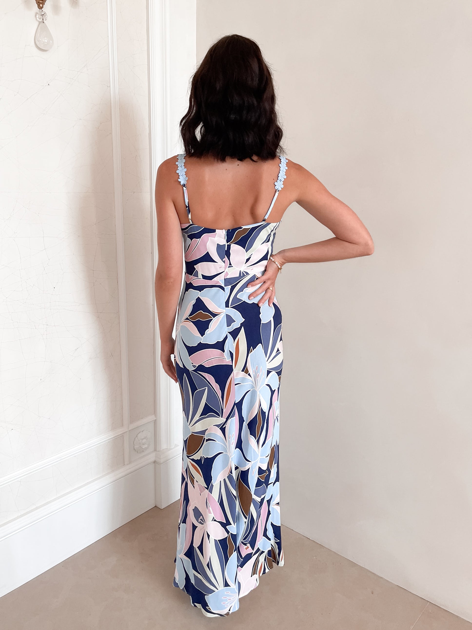 A woman stands smiling indoors against a light-colored wall, wearing a fitted, floor-length dress with a bold floral print in shades of navy blue, light blue, pink, and white. The dress features a deep V-neckline and light blue floral straps. Her dark, wavy hair is styled down, and she accessorizes with a simple gold necklace and rings. She poses confidently with her hands on her hips and is barefoot, adding to the relaxed and elegant vibe of the scene