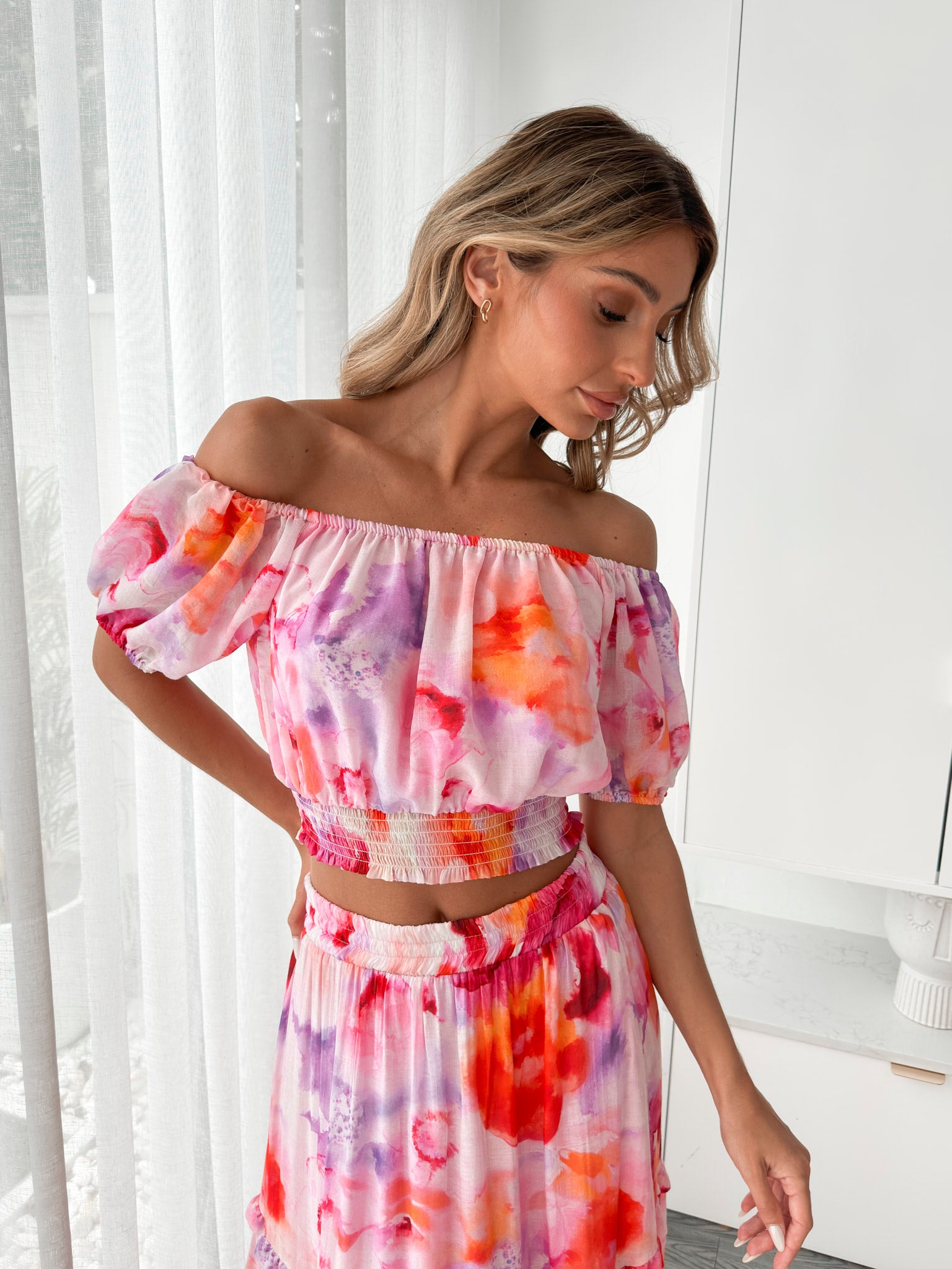 A woman stands indoors in a well-lit, modern room with sheer white curtains and minimalist white cabinetry. She is wearing a colorful off-the-shoulder two-piece outfit consisting of a ruffled crop top and a flowing maxi skirt with a vibrant watercolor floral print in pink, purple, orange, and red hues. Her wavy hair is styled loosely, and she stands gracefully with her hands gently clasped in front, exuding a fresh, summery elegance.