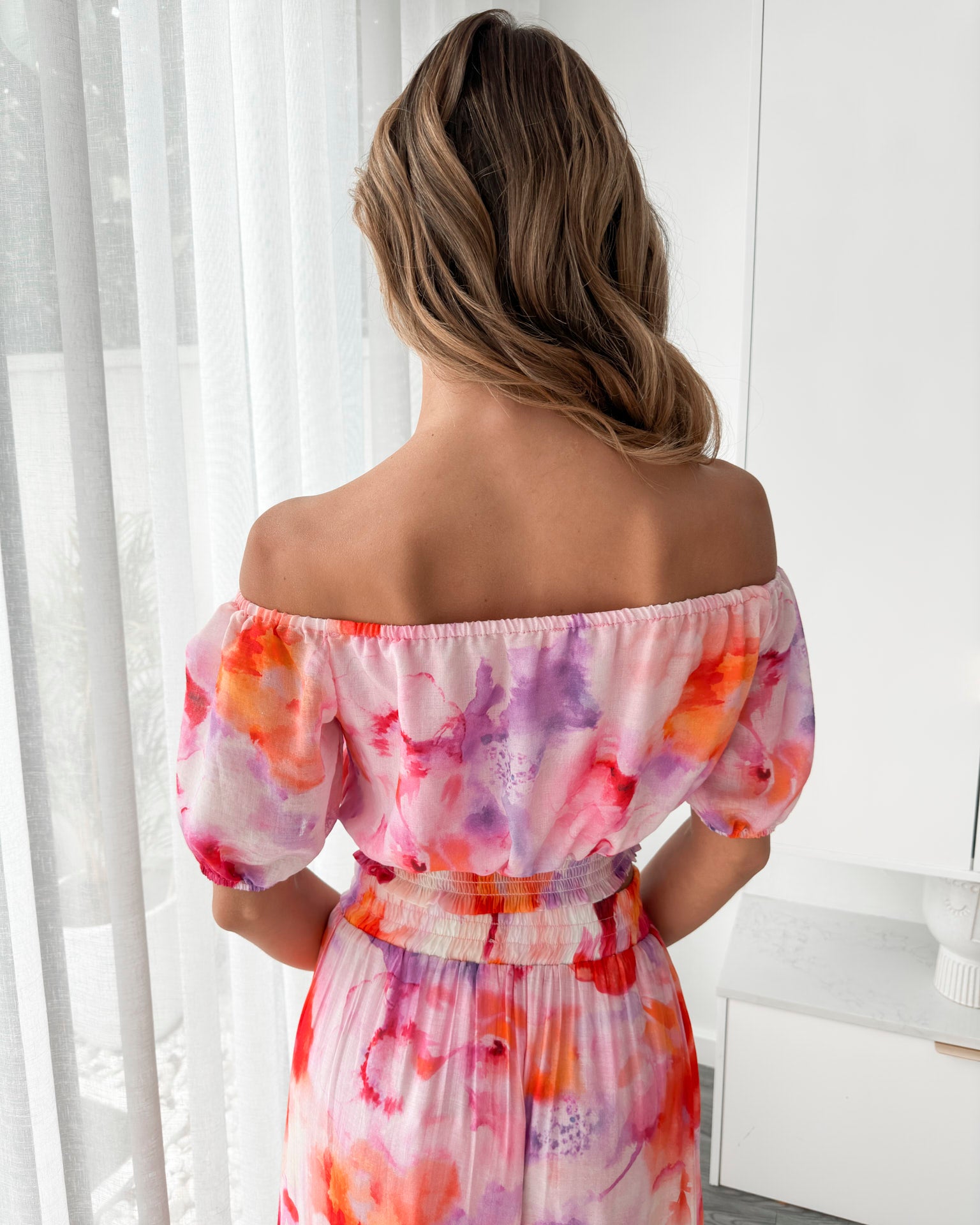 A woman stands indoors in a well-lit, modern room with sheer white curtains and minimalist white cabinetry. She is wearing a colorful off-the-shoulder two-piece outfit consisting of a ruffled crop top and a flowing maxi skirt with a vibrant watercolor floral print in pink, purple, orange, and red hues. Her wavy hair is styled loosely, and she stands gracefully with her hands gently clasped in front, exuding a fresh, summery elegance.