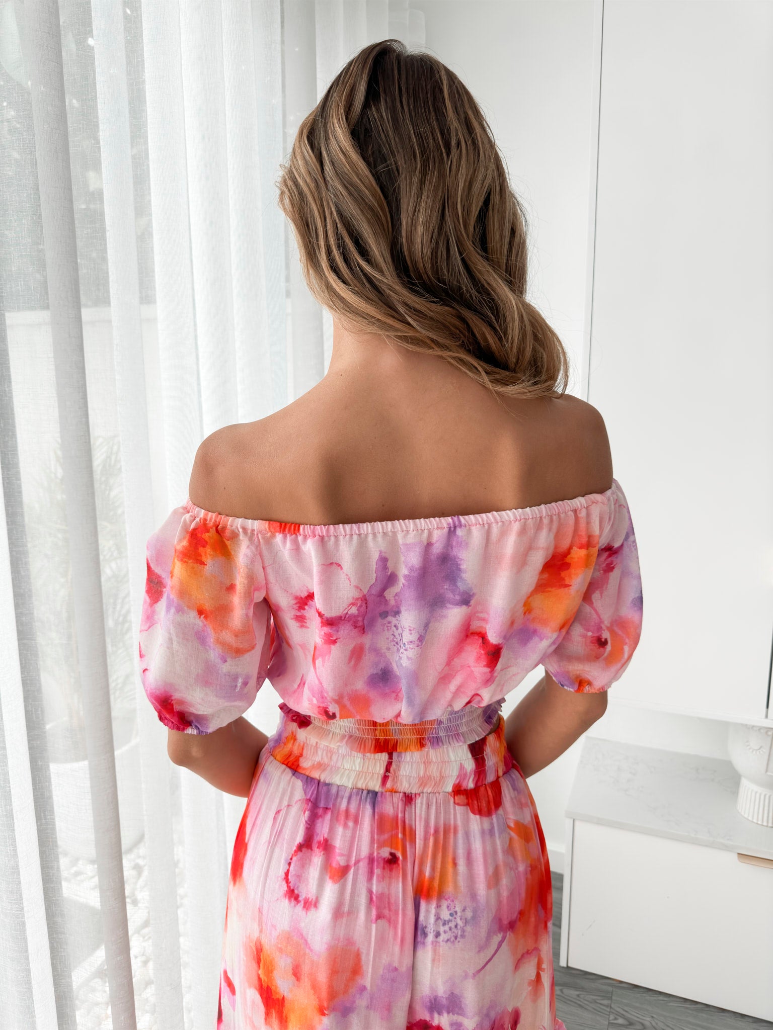 A woman stands indoors in a well-lit, modern room with sheer white curtains and minimalist white cabinetry. She is wearing a colorful off-the-shoulder two-piece outfit consisting of a ruffled crop top and a flowing maxi skirt with a vibrant watercolor floral print in pink, purple, orange, and red hues. Her wavy hair is styled loosely, and she stands gracefully with her hands gently clasped in front, exuding a fresh, summery elegance.