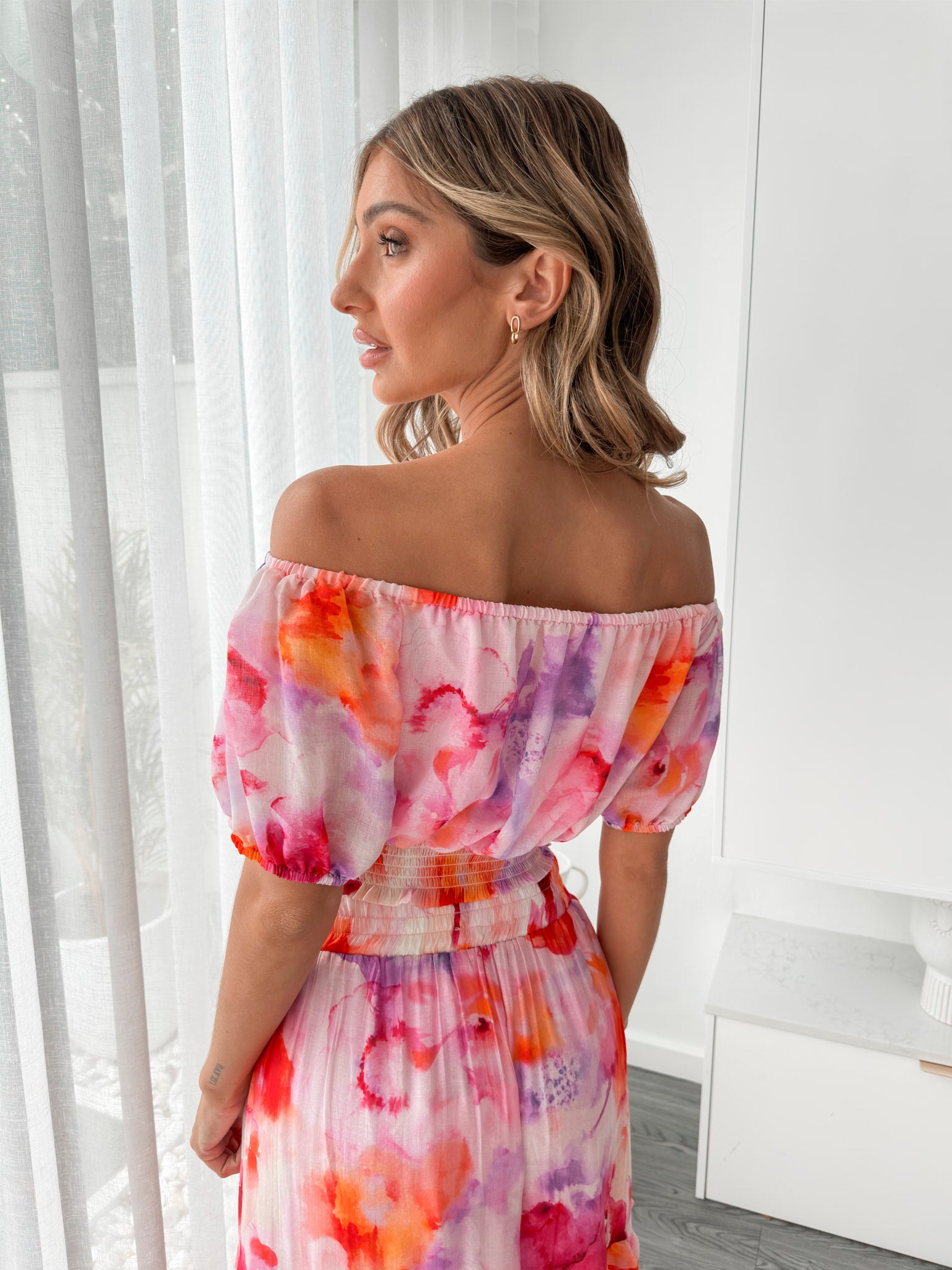 A woman stands indoors in a well-lit, modern room with sheer white curtains and minimalist white cabinetry. She is wearing a colorful off-the-shoulder two-piece outfit consisting of a ruffled crop top and a flowing maxi skirt with a vibrant watercolor floral print in pink, purple, orange, and red hues. Her wavy hair is styled loosely, and she stands gracefully with her hands gently clasped in front, exuding a fresh, summery elegance.