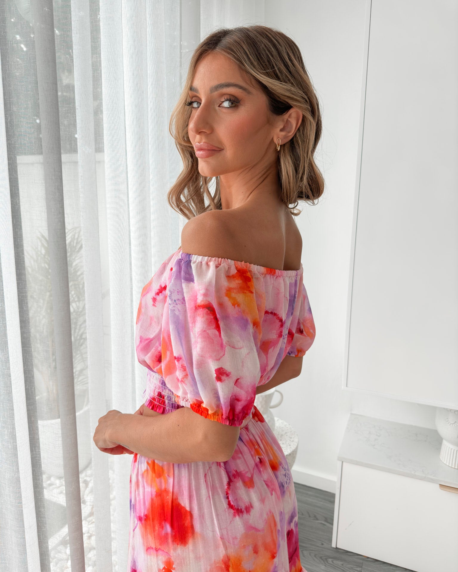 A woman stands indoors in a well-lit, modern room with sheer white curtains and minimalist white cabinetry. She is wearing a colorful off-the-shoulder two-piece outfit consisting of a ruffled crop top and a flowing maxi skirt with a vibrant watercolor floral print in pink, purple, orange, and red hues. Her wavy hair is styled loosely, and she stands gracefully with her hands gently clasped in front, exuding a fresh, summery elegance.