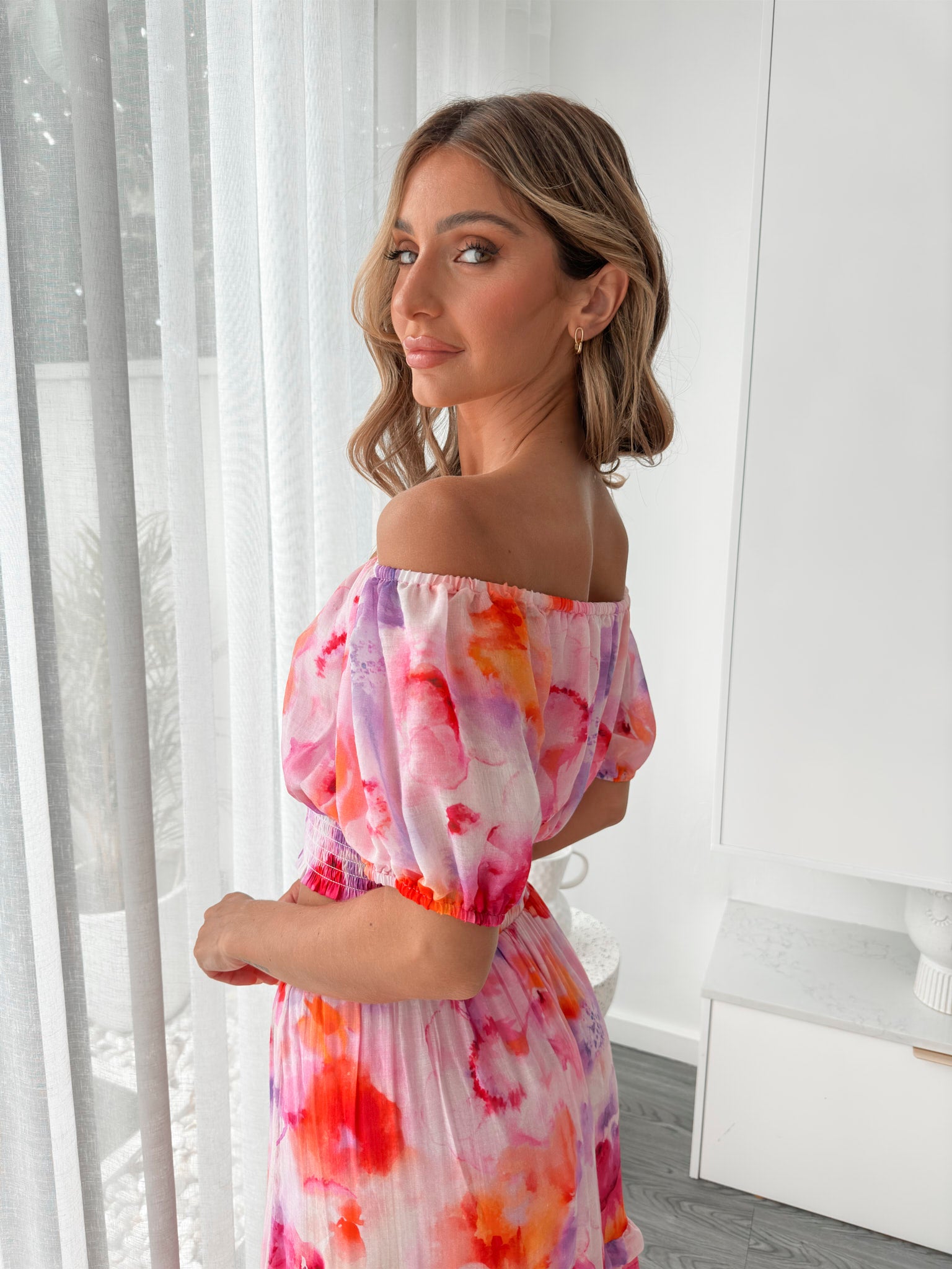 A woman stands indoors in a well-lit, modern room with sheer white curtains and minimalist white cabinetry. She is wearing a colorful off-the-shoulder two-piece outfit consisting of a ruffled crop top and a flowing maxi skirt with a vibrant watercolor floral print in pink, purple, orange, and red hues. Her wavy hair is styled loosely, and she stands gracefully with her hands gently clasped in front, exuding a fresh, summery elegance.