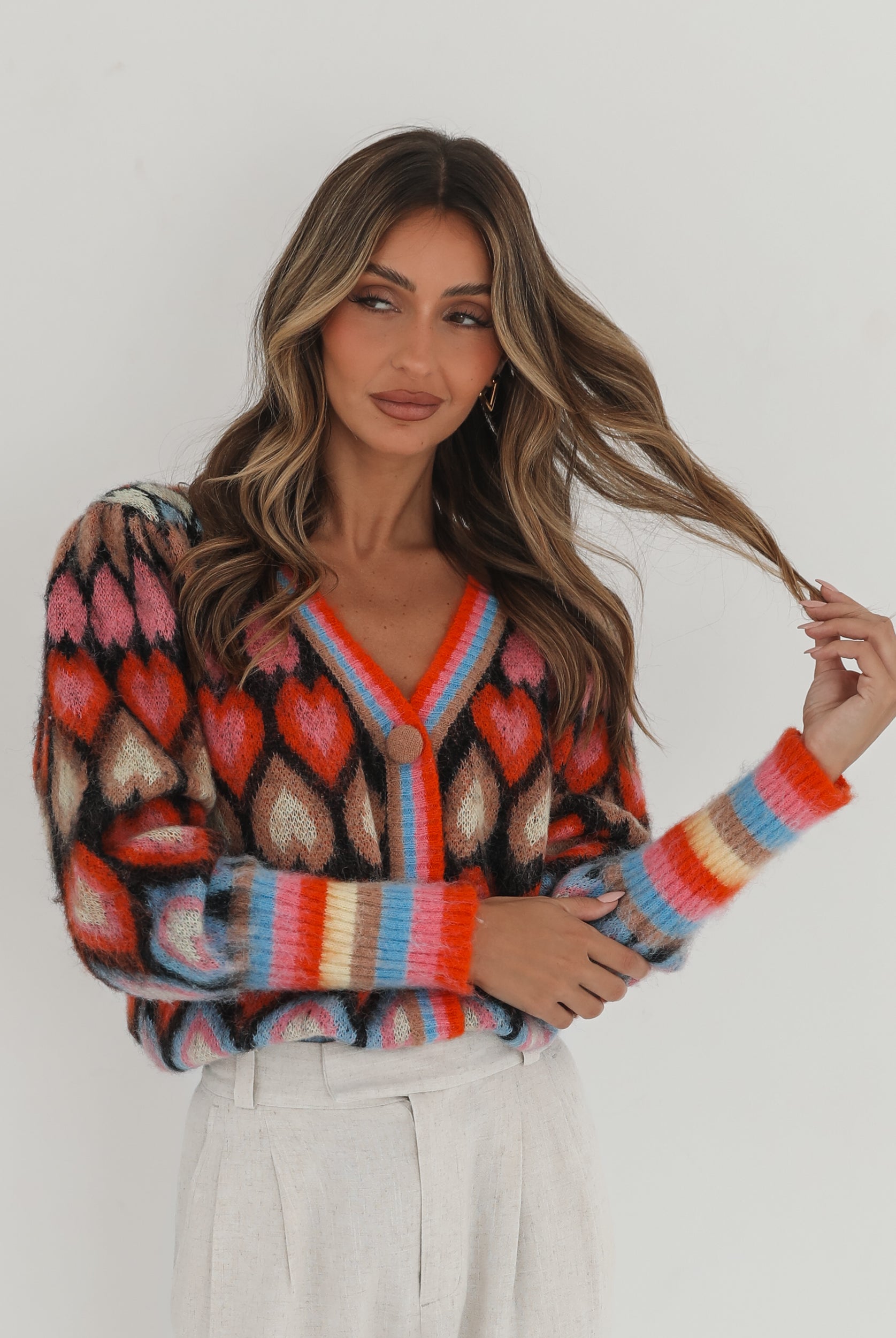 A woman poses against a plain white background wearing a colorful, patterned cardigan with heart shapes and bold stripes in red, pink, blue, and orange tones. She pairs it with high-waisted, light beige trousers and has long, wavy brown hair with blonde highlights. She is looking to the side and gently touching her hair with one hand.