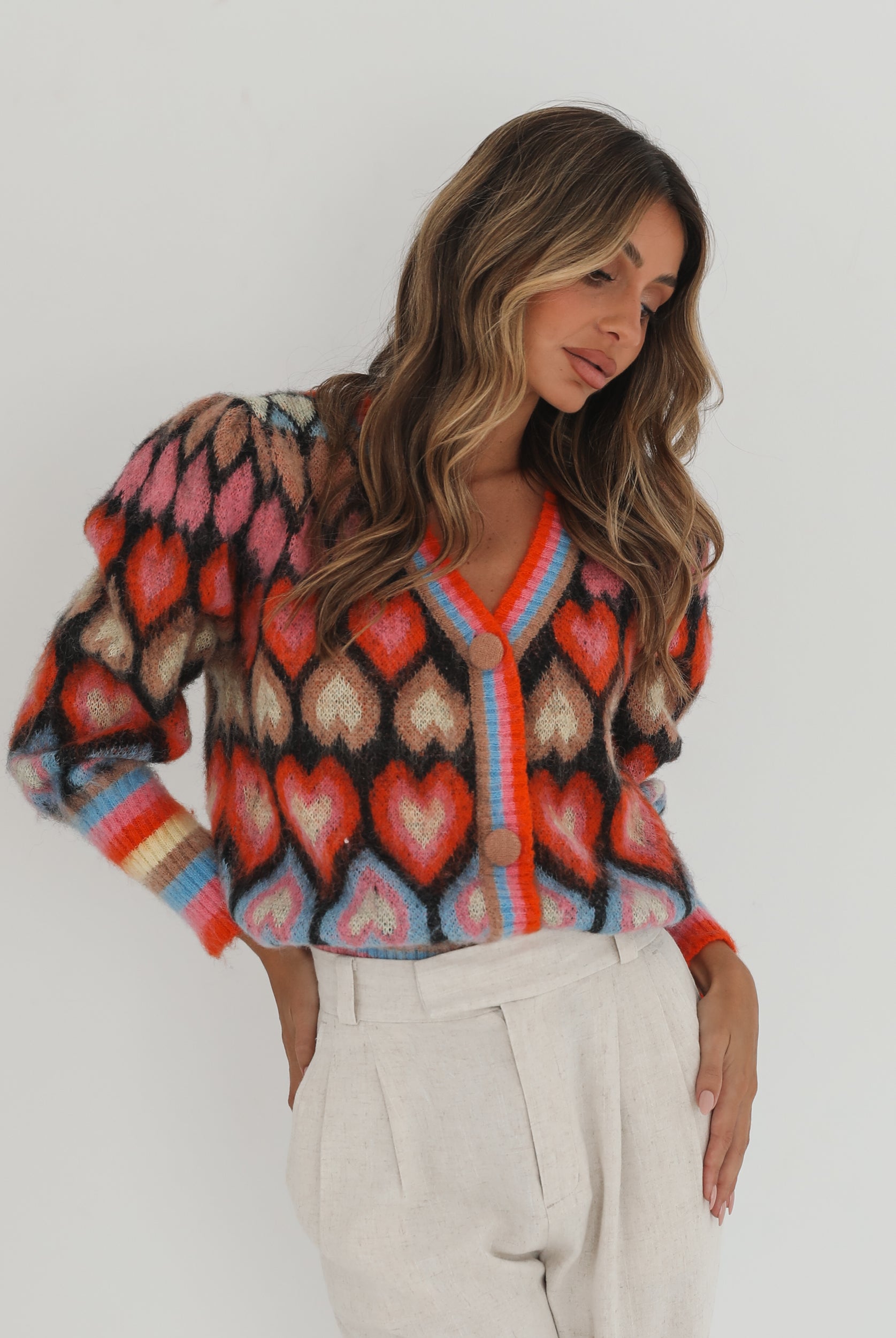 A woman poses against a plain white background wearing a colorful, patterned cardigan with heart shapes and bold stripes in red, pink, blue, and orange tones. She pairs it with high-waisted, light beige trousers and has long, wavy brown hair with blonde highlights. She is looking to the side and gently touching her hair with one hand.