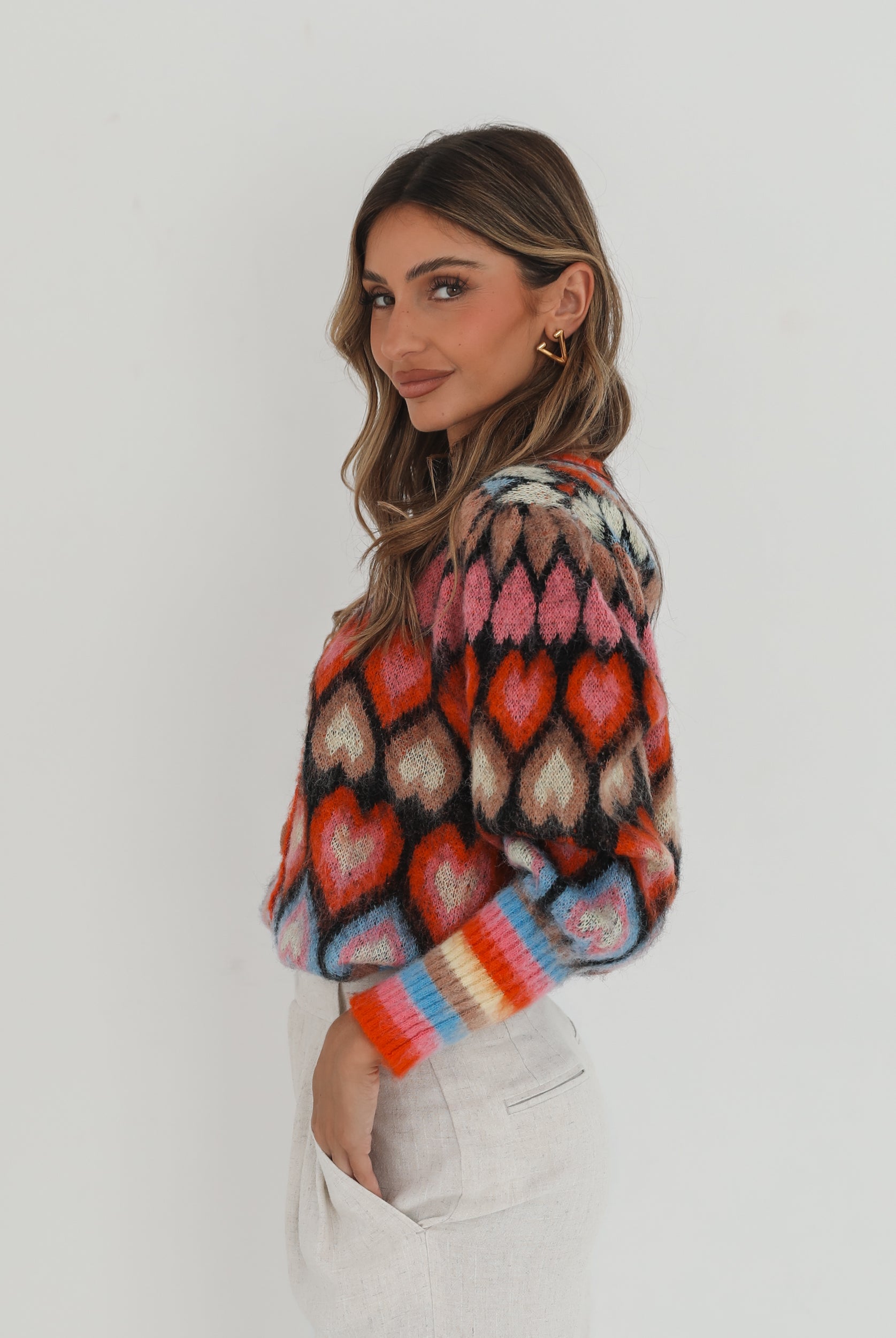 A woman poses against a plain white background wearing a colorful, patterned cardigan with heart shapes and bold stripes in red, pink, blue, and orange tones. She pairs it with high-waisted, light beige trousers and has long, wavy brown hair with blonde highlights. She is looking to the side and gently touching her hair with one hand.