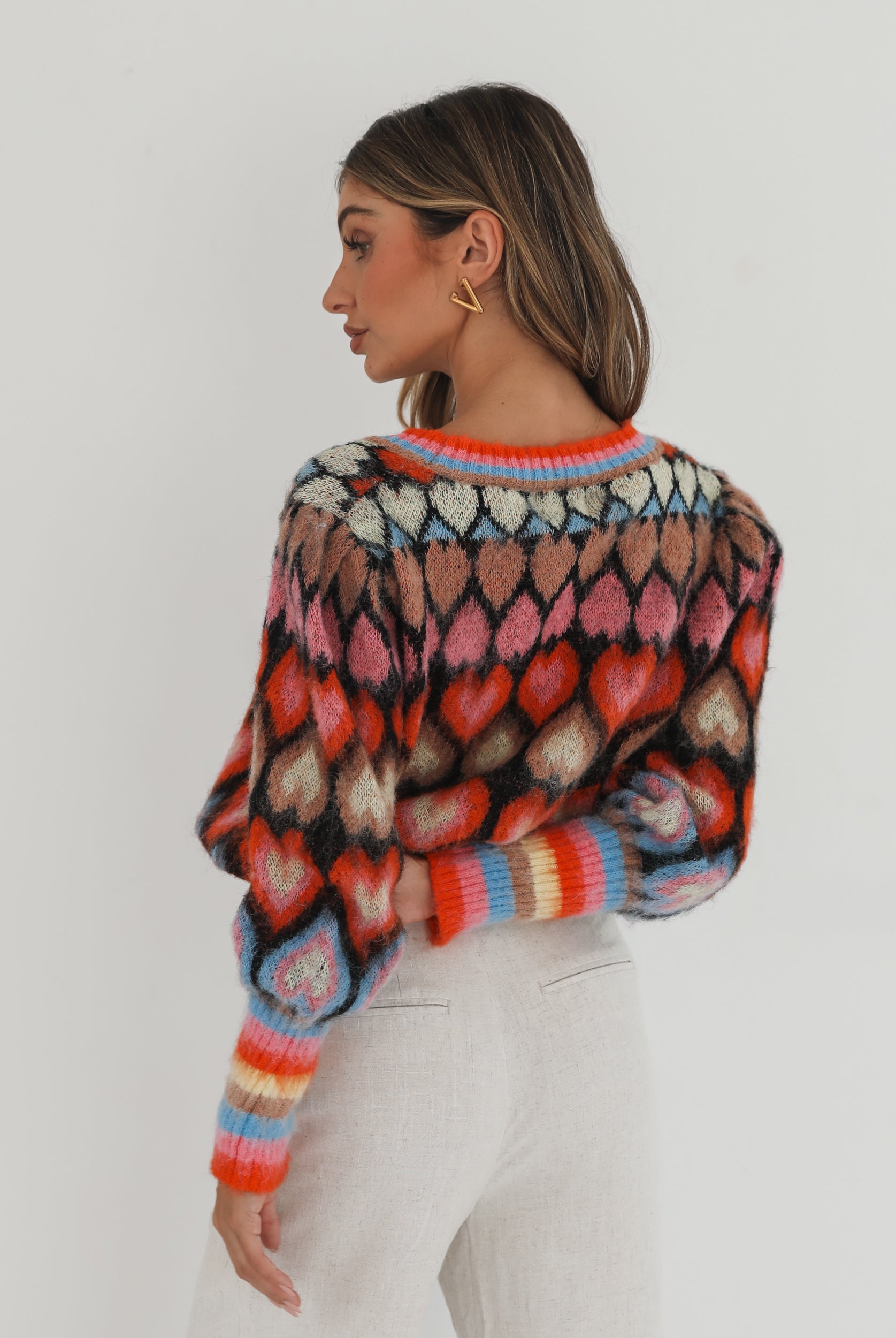 A woman poses against a plain white background wearing a colorful, patterned cardigan with heart shapes and bold stripes in red, pink, blue, and orange tones. She pairs it with high-waisted, light beige trousers and has long, wavy brown hair with blonde highlights. She is looking to the side and gently touching her hair with one hand.