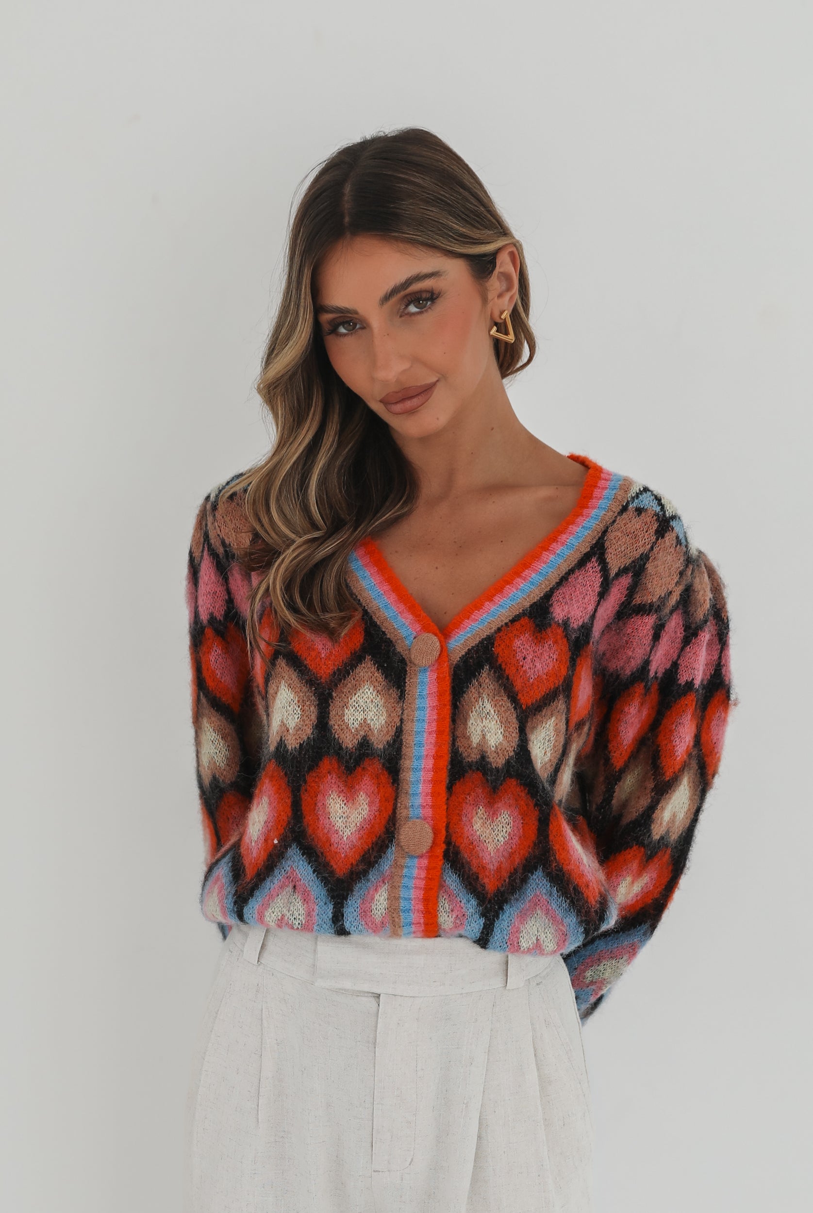 A woman poses against a plain white background wearing a colorful, patterned cardigan with heart shapes and bold stripes in red, pink, blue, and orange tones. She pairs it with high-waisted, light beige trousers and has long, wavy brown hair with blonde highlights. She is looking to the side and gently touching her hair with one hand.