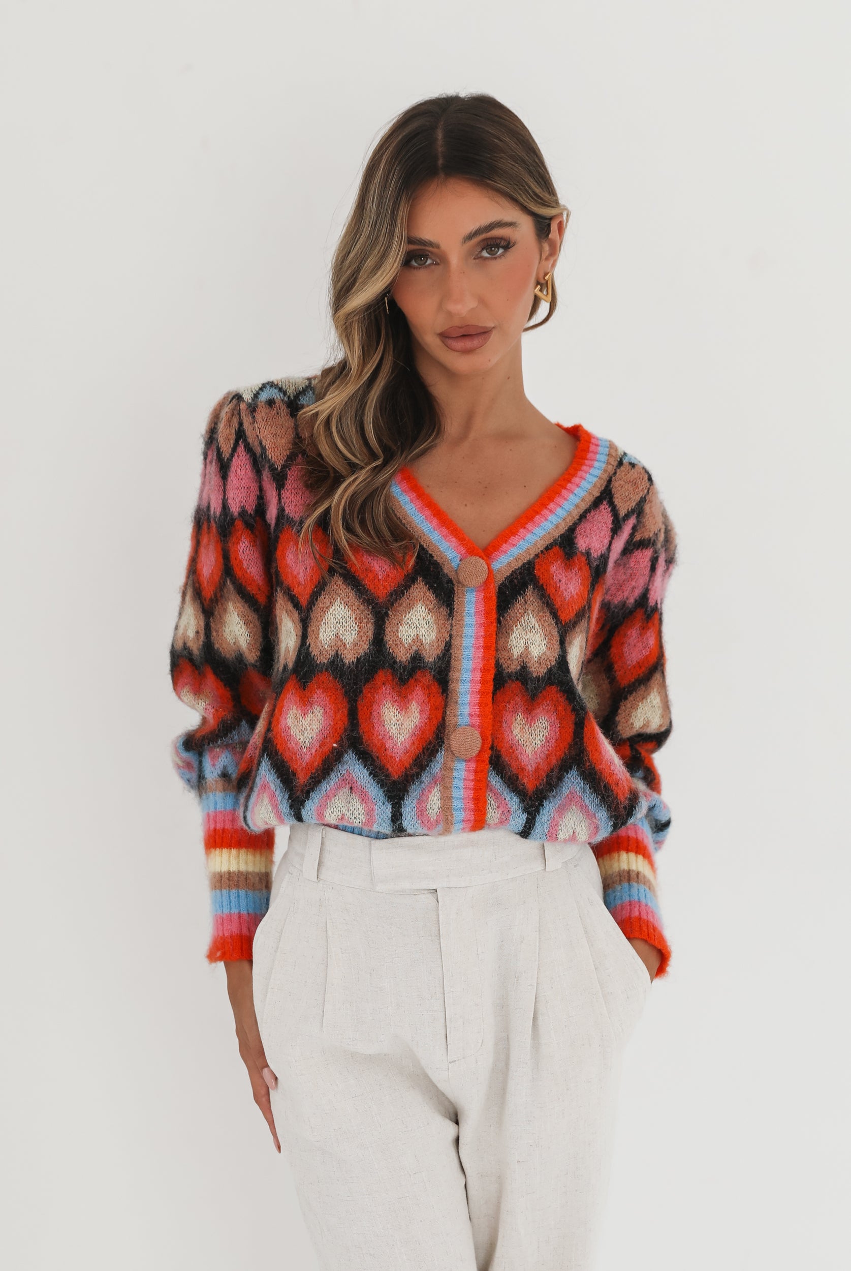 A woman poses against a plain white background wearing a colorful, patterned cardigan with heart shapes and bold stripes in red, pink, blue, and orange tones. She pairs it with high-waisted, light beige trousers and has long, wavy brown hair with blonde highlights. She is looking to the side and gently touching her hair with one hand.