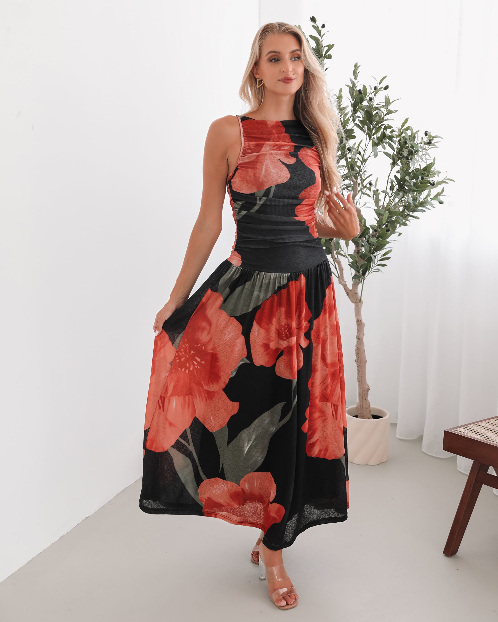 A woman stands indoors near a potted plant, wearing a sleeveless black maxi dress with a bold red floral print. Her long blonde hair is styled in loose waves, and she accessorizes with gold hoop earrings and clear-heeled sandals. She smiles softly while posing with her hands gently holding the sides of her dress.
