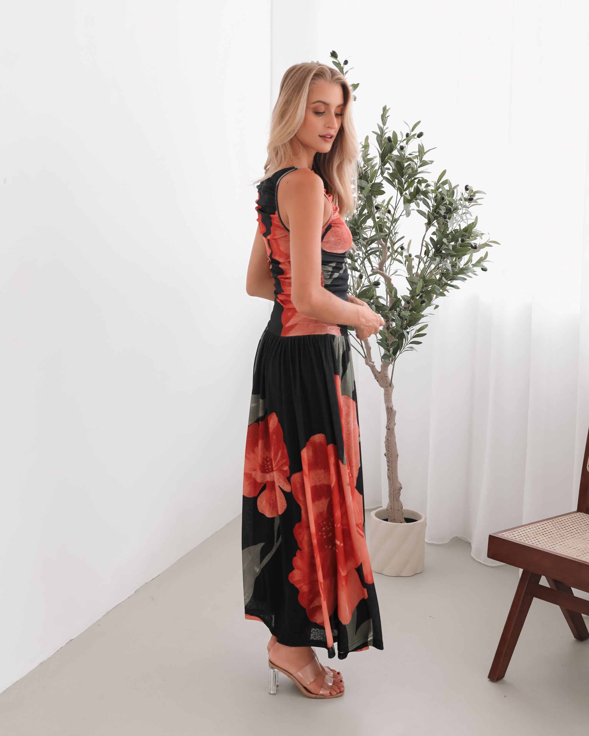 A woman stands indoors near a potted plant, wearing a sleeveless black maxi dress with a bold red floral print. Her long blonde hair is styled in loose waves, and she accessorizes with gold hoop earrings and clear-heeled sandals. She smiles softly while posing with her hands gently holding the sides of her dress.