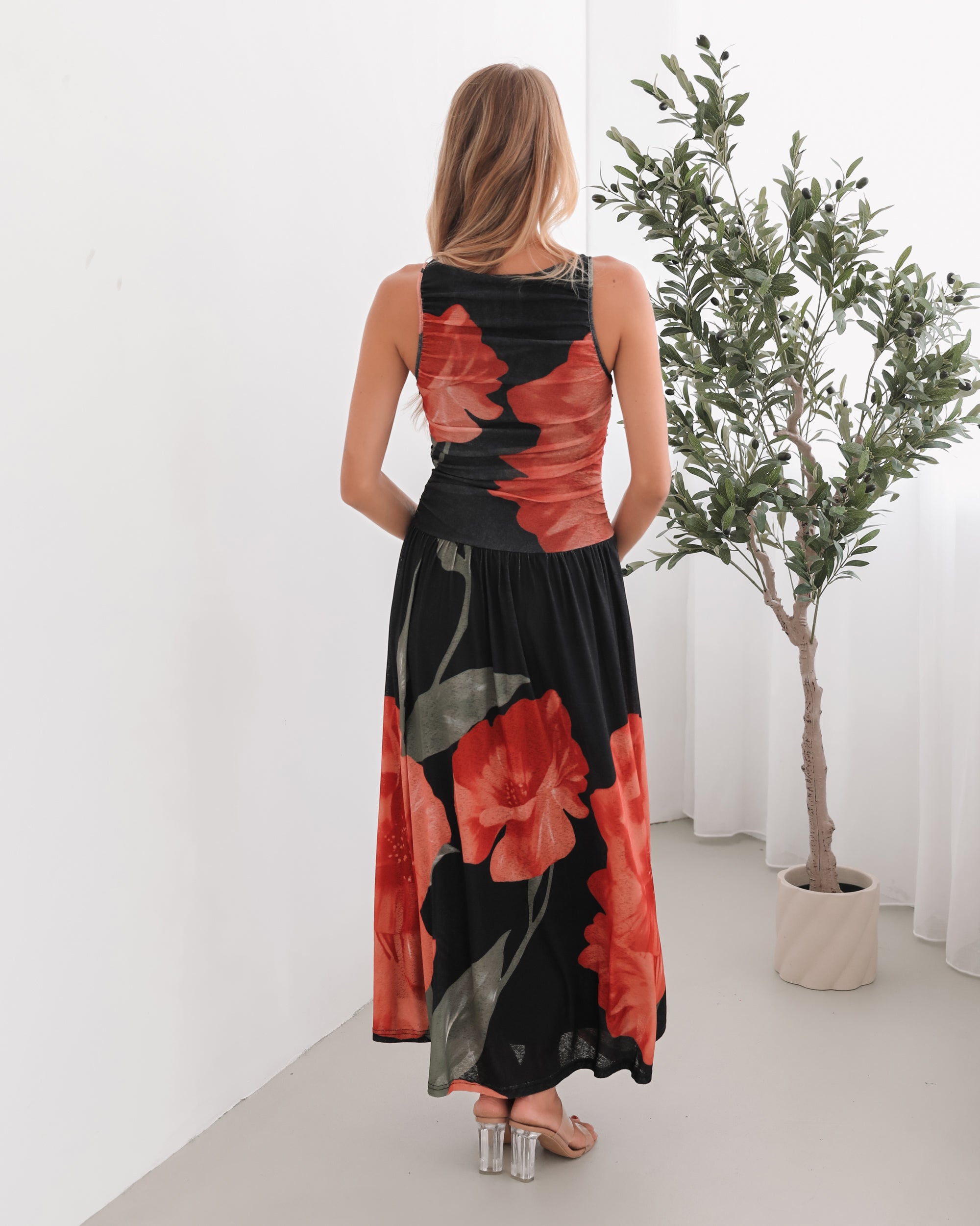 A woman stands indoors near a potted plant, wearing a sleeveless black maxi dress with a bold red floral print. Her long blonde hair is styled in loose waves, and she accessorizes with gold hoop earrings and clear-heeled sandals. She smiles softly while posing with her hands gently holding the sides of her dress.