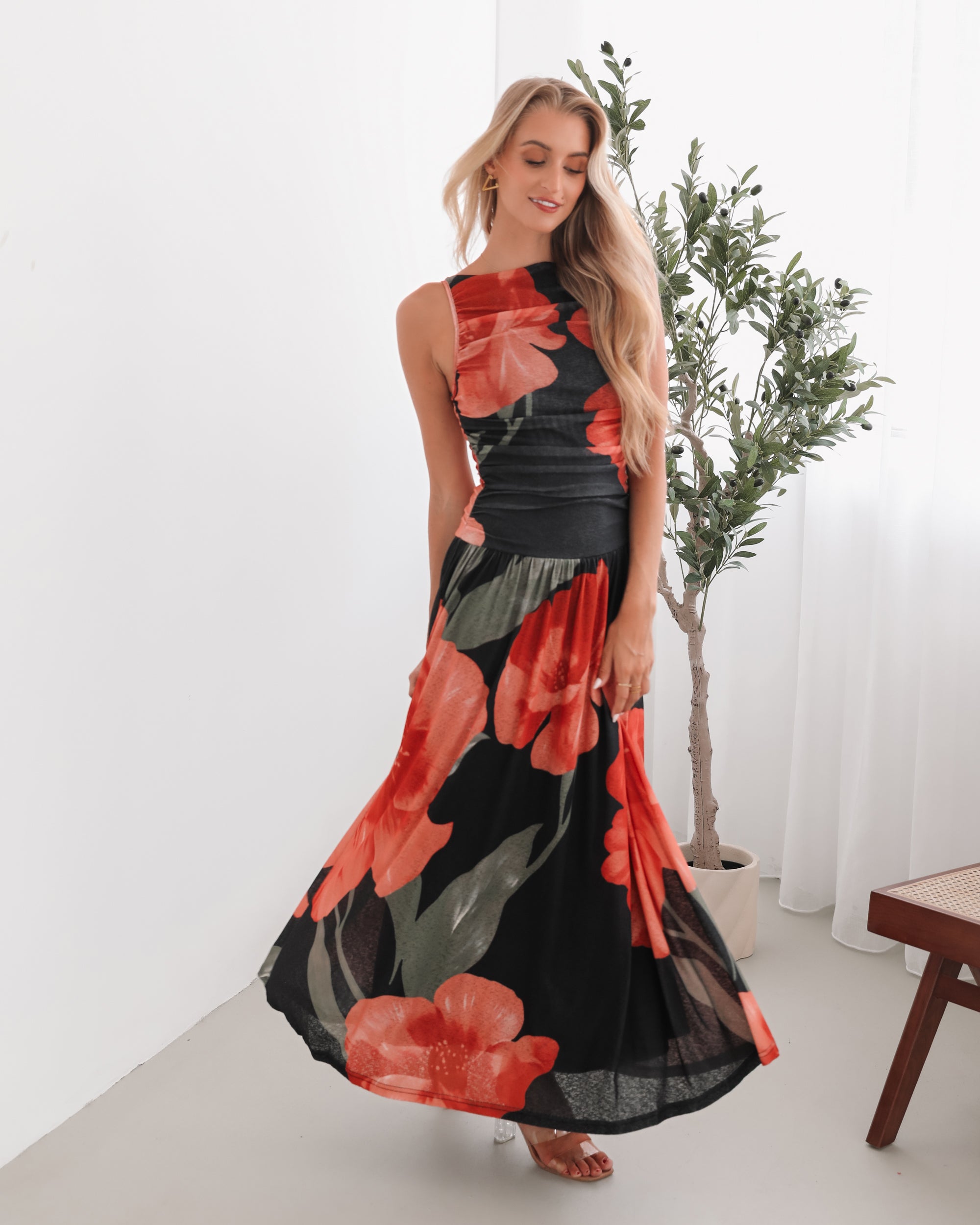 A woman stands indoors near a potted plant, wearing a sleeveless black maxi dress with a bold red floral print. Her long blonde hair is styled in loose waves, and she accessorizes with gold hoop earrings and clear-heeled sandals. She smiles softly while posing with her hands gently holding the sides of her dress.