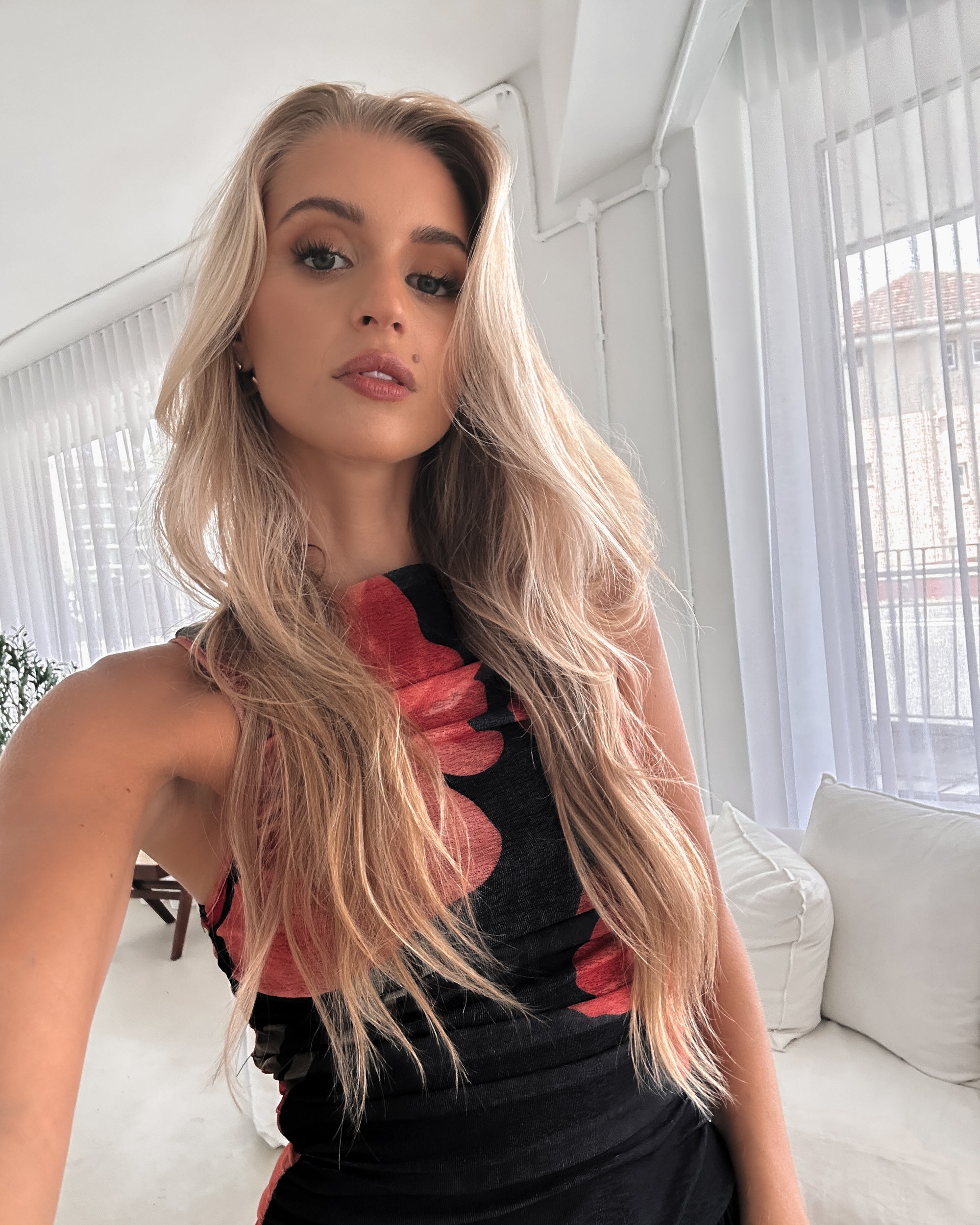 A woman stands indoors near a potted plant, wearing a sleeveless black maxi dress with a bold red floral print. Her long blonde hair is styled in loose waves, and she accessorizes with gold hoop earrings and clear-heeled sandals. She smiles softly while posing with her hands gently holding the sides of her dress.