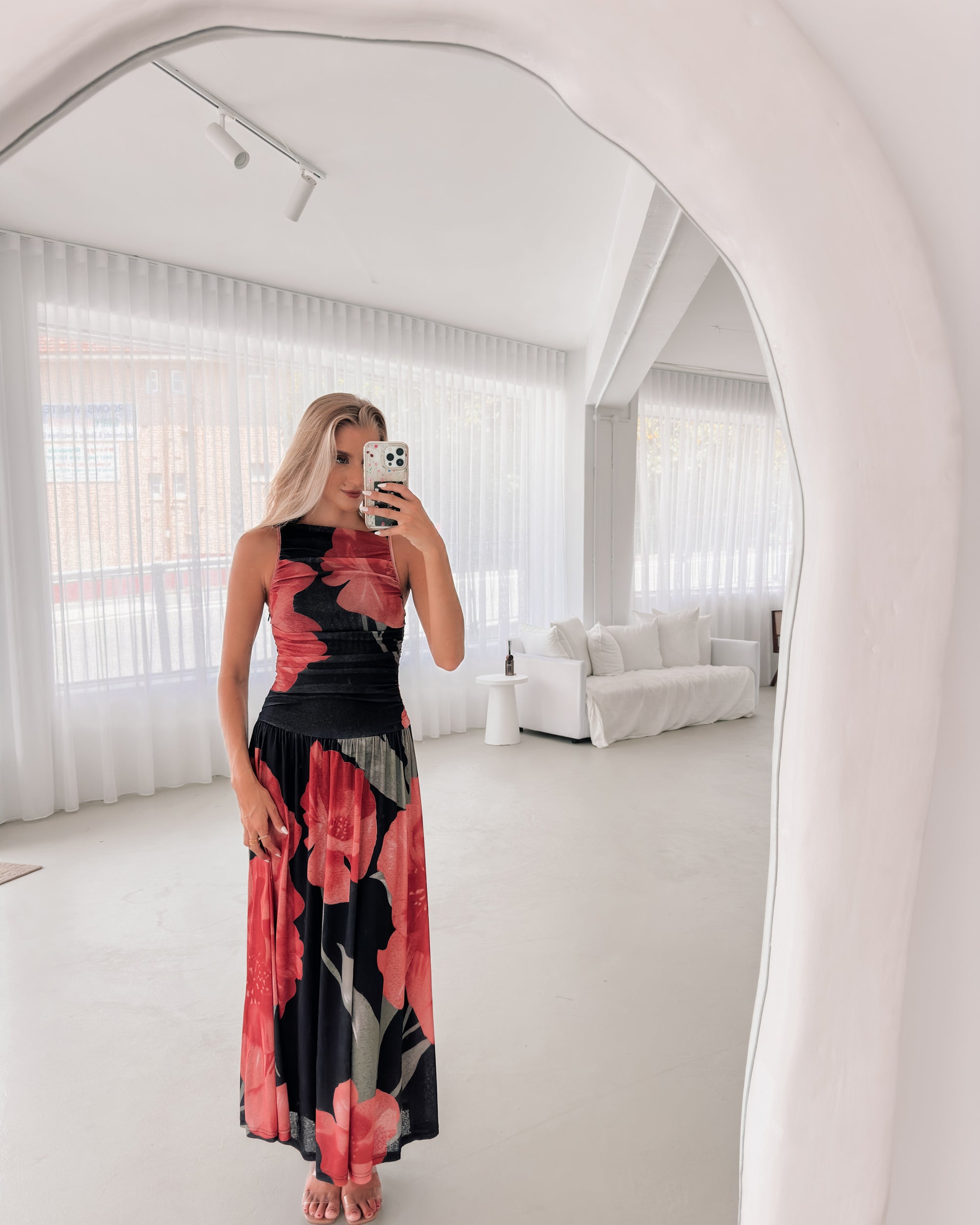 A woman stands indoors near a potted plant, wearing a sleeveless black maxi dress with a bold red floral print. Her long blonde hair is styled in loose waves, and she accessorizes with gold hoop earrings and clear-heeled sandals. She smiles softly while posing with her hands gently holding the sides of her dress.