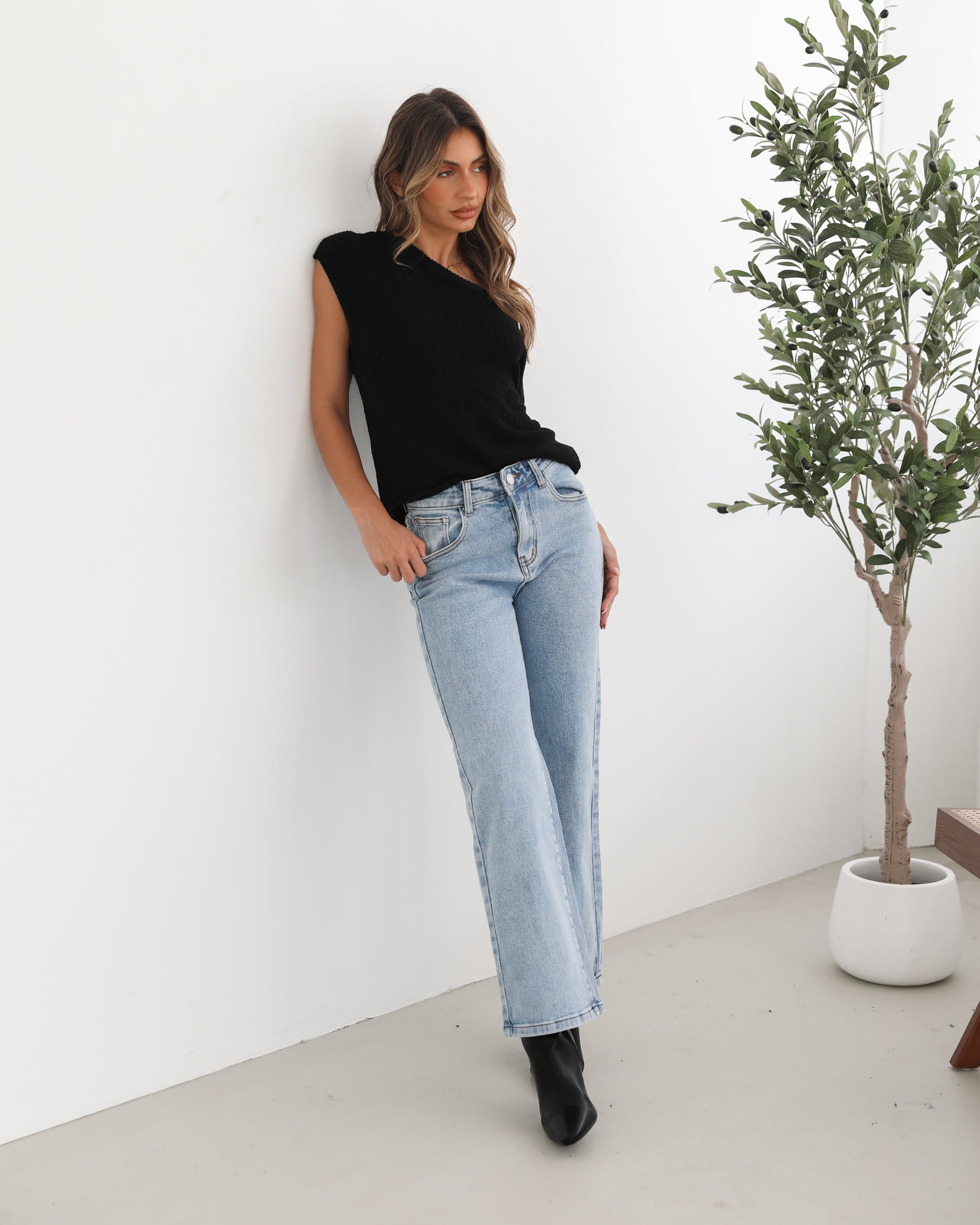 Woman wearing light-wash flared jeans and black heeled ankle boots, paired with a sleeveless black top, standing indoors near a potted plant and a wooden chair.
