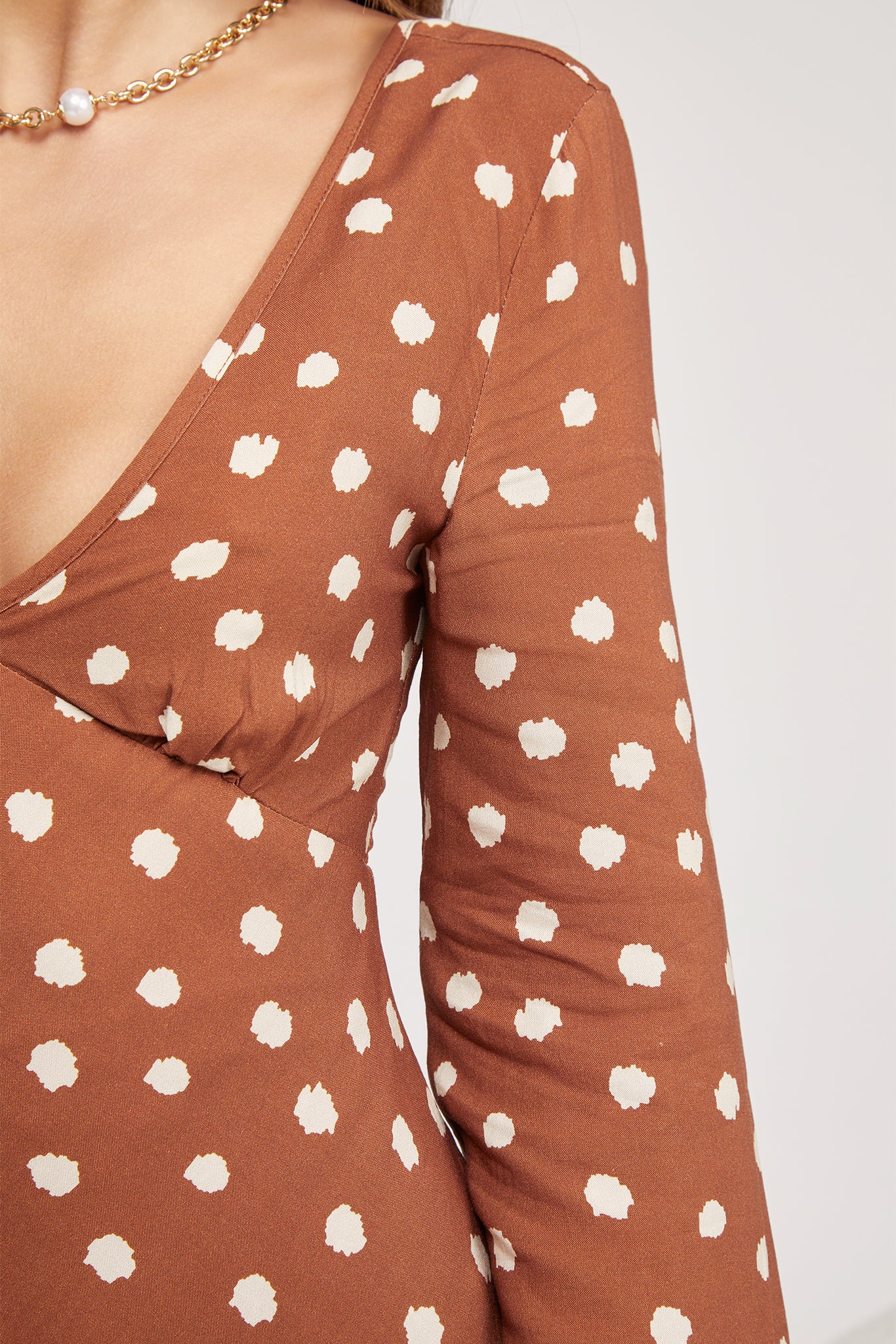 Woman wearing a brown long-sleeve mini dress with white polka dots and flared cuffs, paired with light beige ankle boots, standing in a modern, minimalist room with a white chair, a small shelf, and a textured rug.