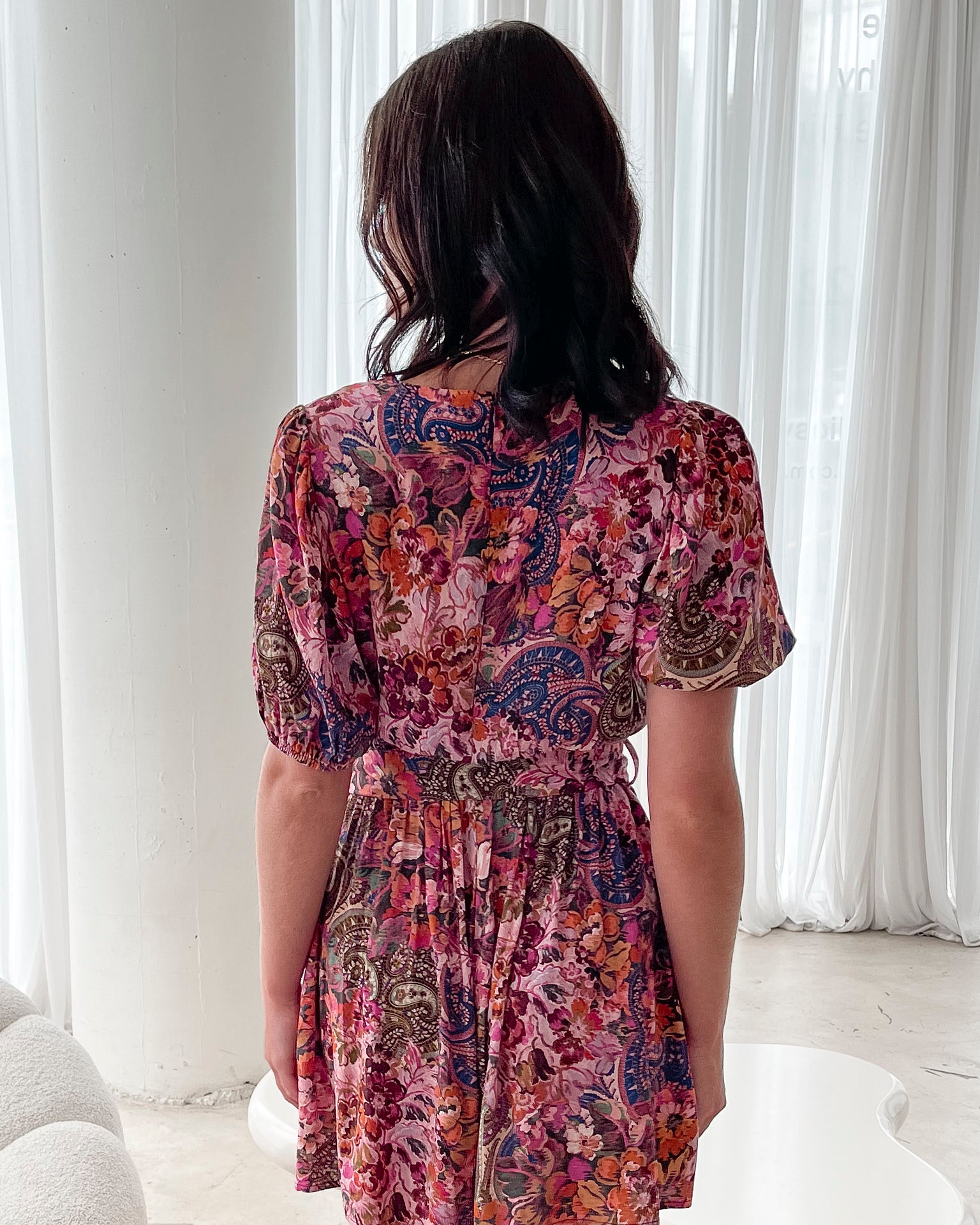 Woman wearing a short, colorful floral and paisley-patterned dress with a deep V-neckline trimmed in lace, puff sleeves, and a belted waist, standing indoors in front of sheer white curtains.