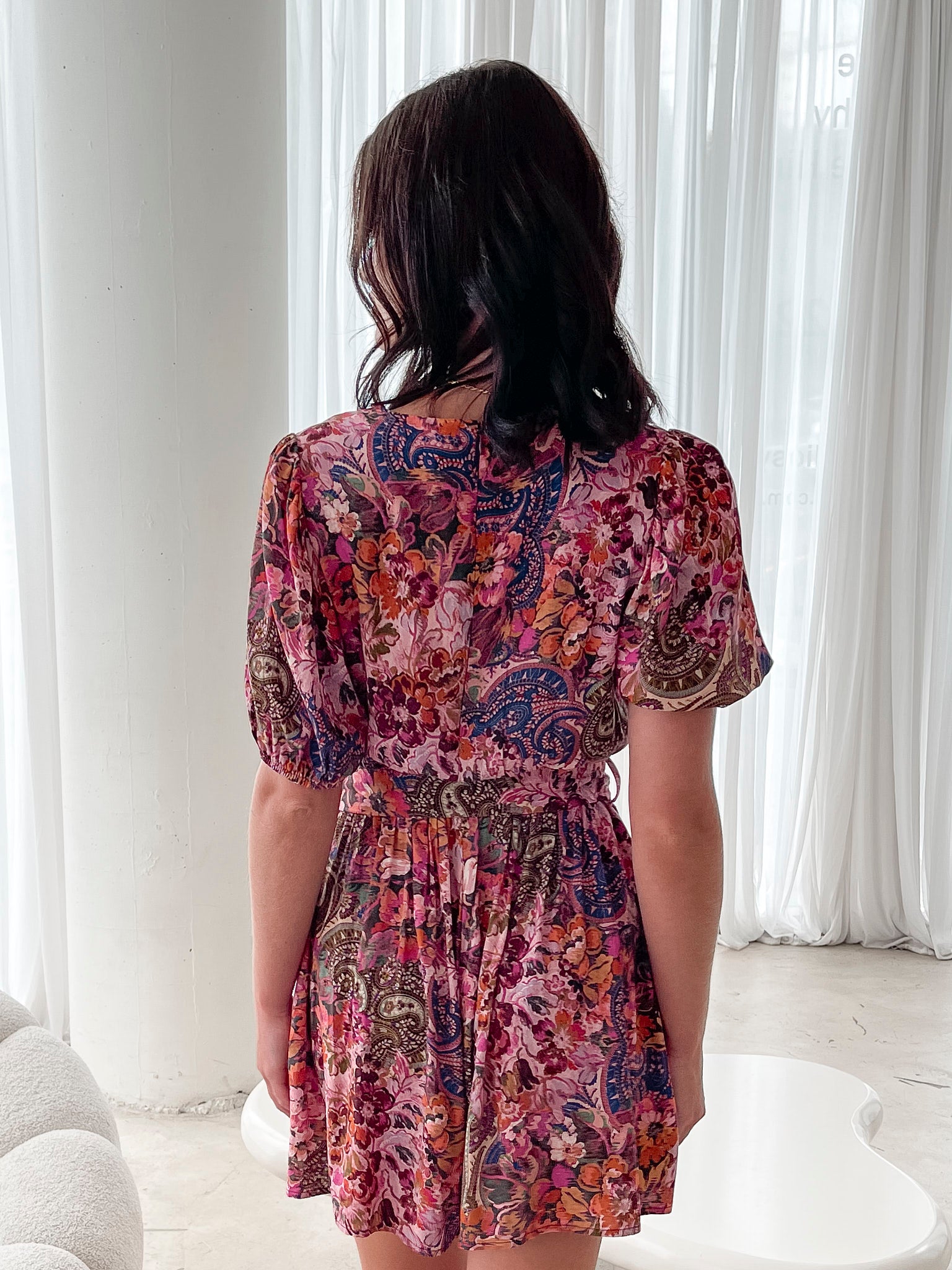 Woman wearing a short, colorful floral and paisley-patterned dress with a deep V-neckline trimmed in lace, puff sleeves, and a belted waist, standing indoors in front of sheer white curtains.