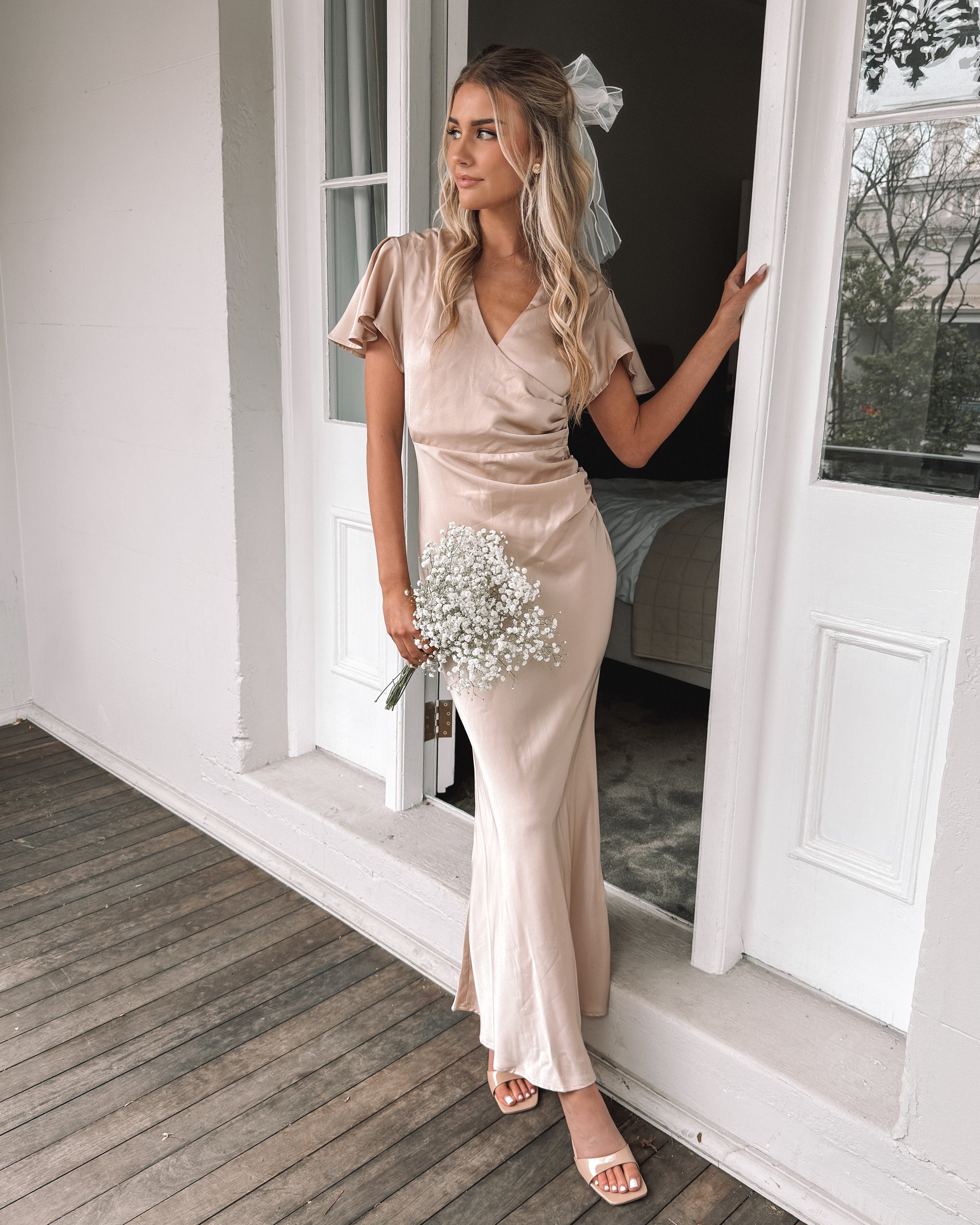 Graceful woman in a beige dress holding a bouquet, suited for wedding celebrations, high-society gatherings, charity dinners, luxury banquets, and elegant affairs, standing in a doorway.