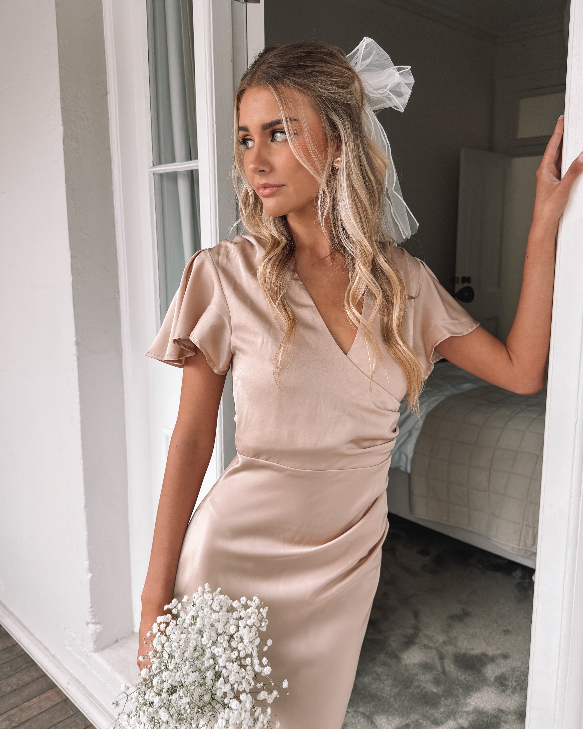 Graceful woman in a beige dress holding a bouquet, suited for wedding celebrations, high-society gatherings, charity dinners, luxury banquets, and elegant affairs, standing in a doorway.