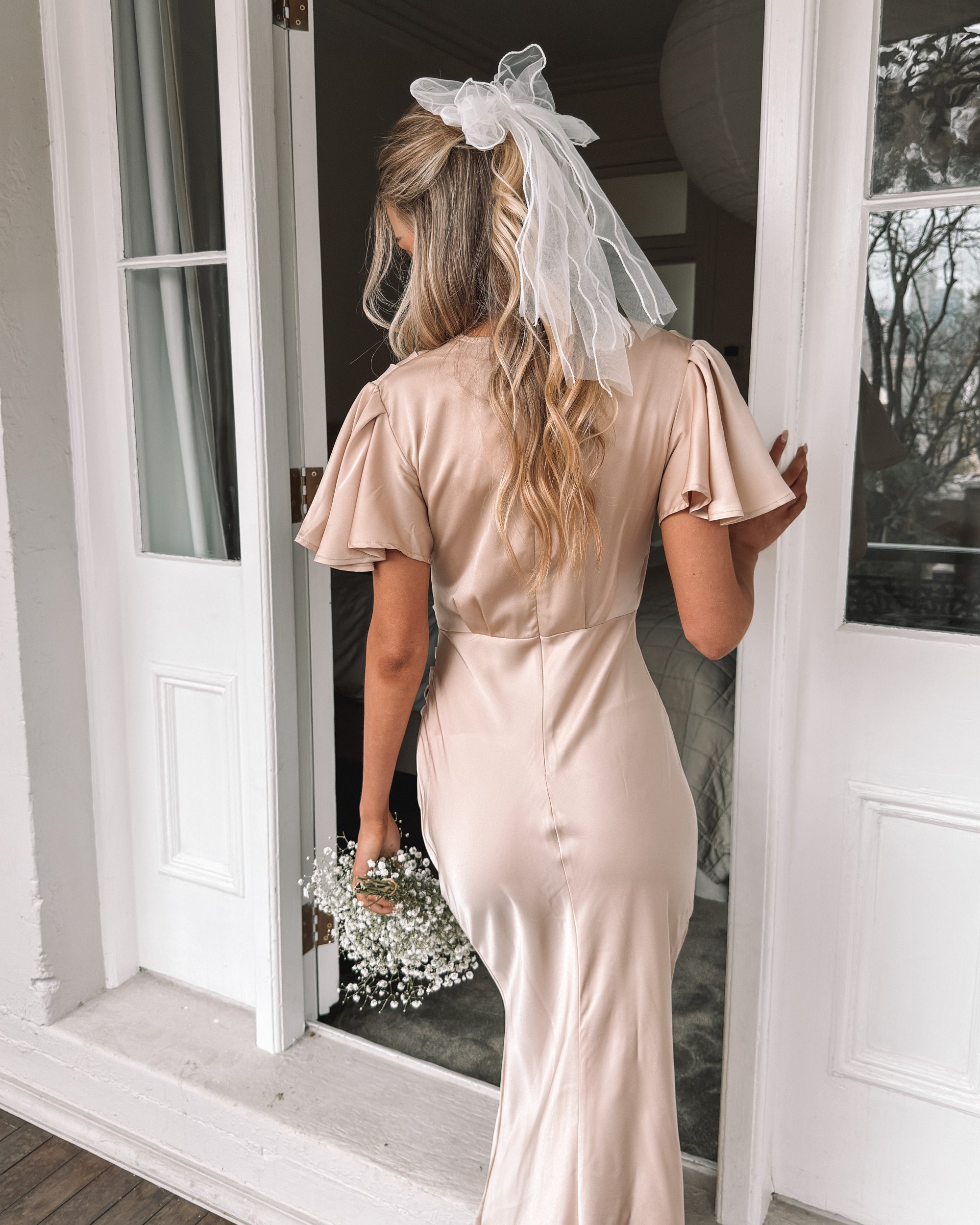 Graceful woman in a beige dress holding a bouquet, suited for wedding celebrations, high-society gatherings, charity dinners, luxury banquets, and elegant affairs, standing in a doorway.