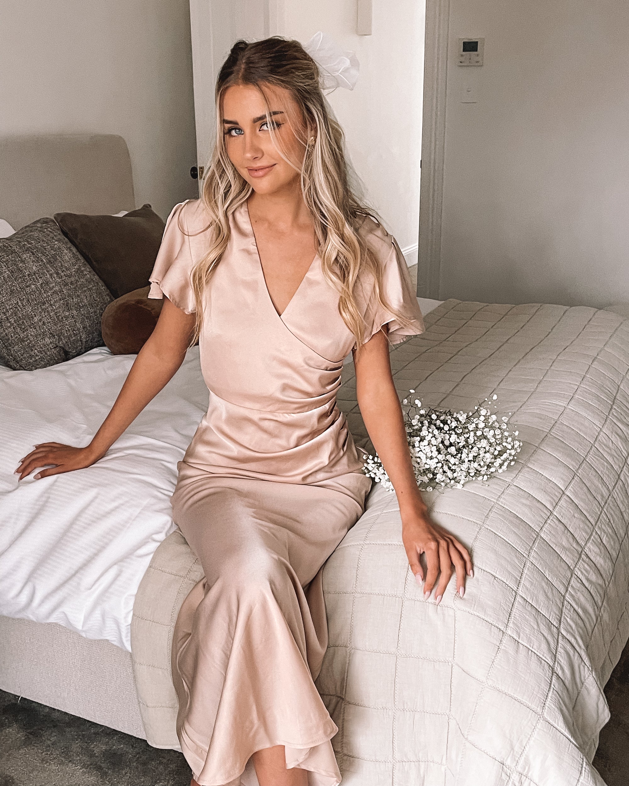 Graceful woman in a beige dress holding a bouquet, suited for wedding celebrations, high-society gatherings, charity dinners, luxury banquets, and elegant affairs, standing in a doorway.
