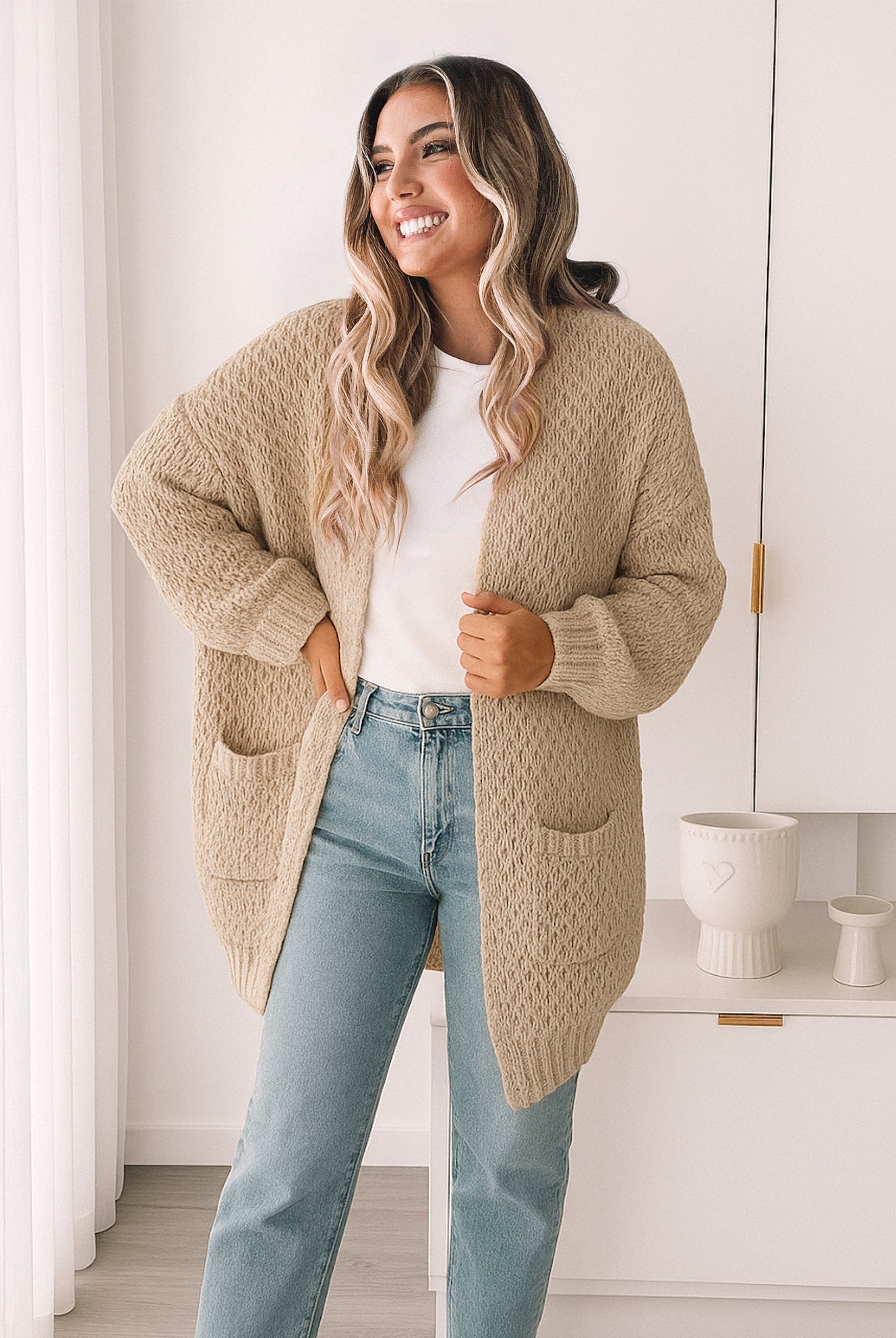 Woman wearing a beige cardigan over a white shirt and light blue jeans in a bright room.