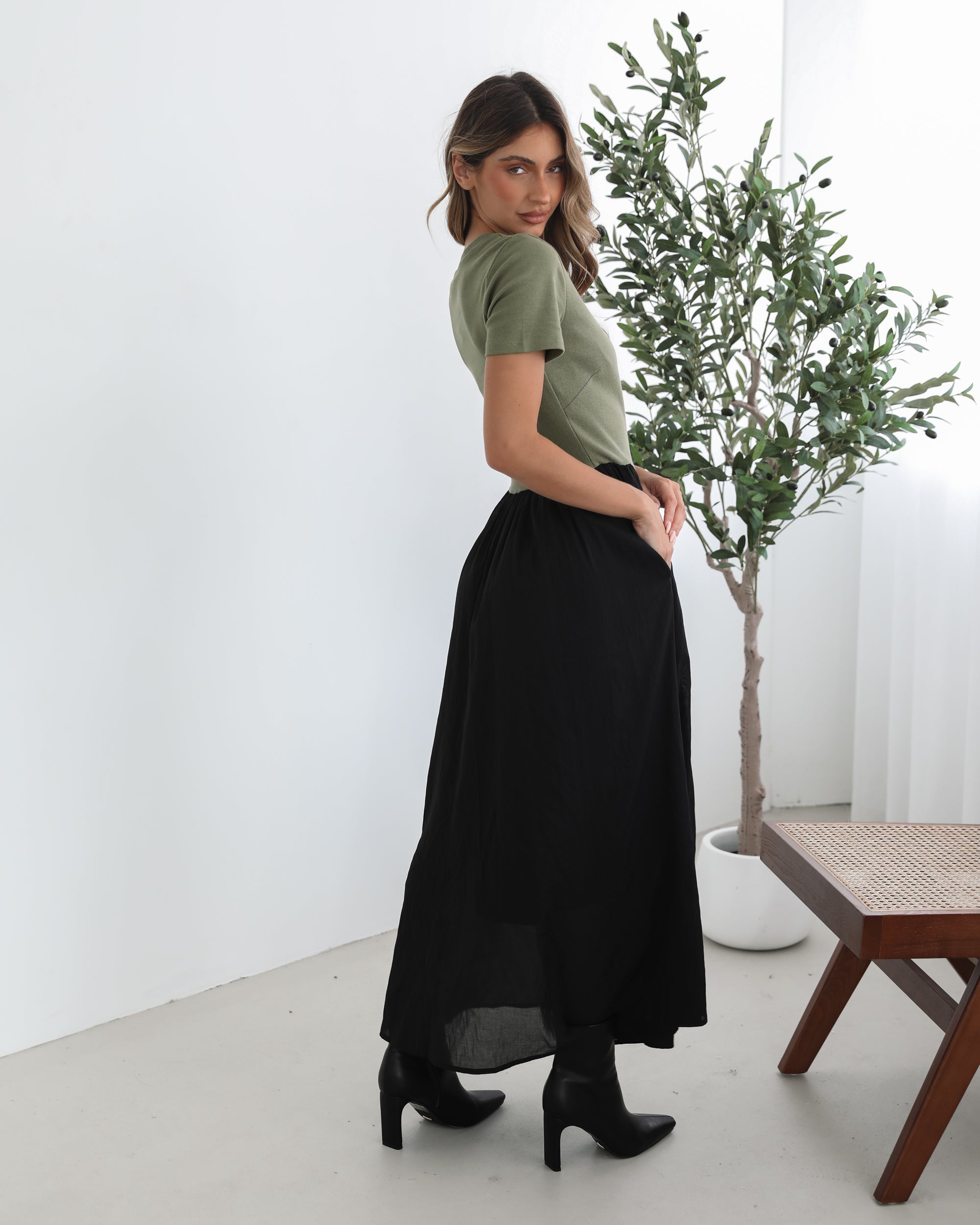 woman wearing a short sleeve round neck dress with green top and black flowy skirt paired with black boots in a white background studio