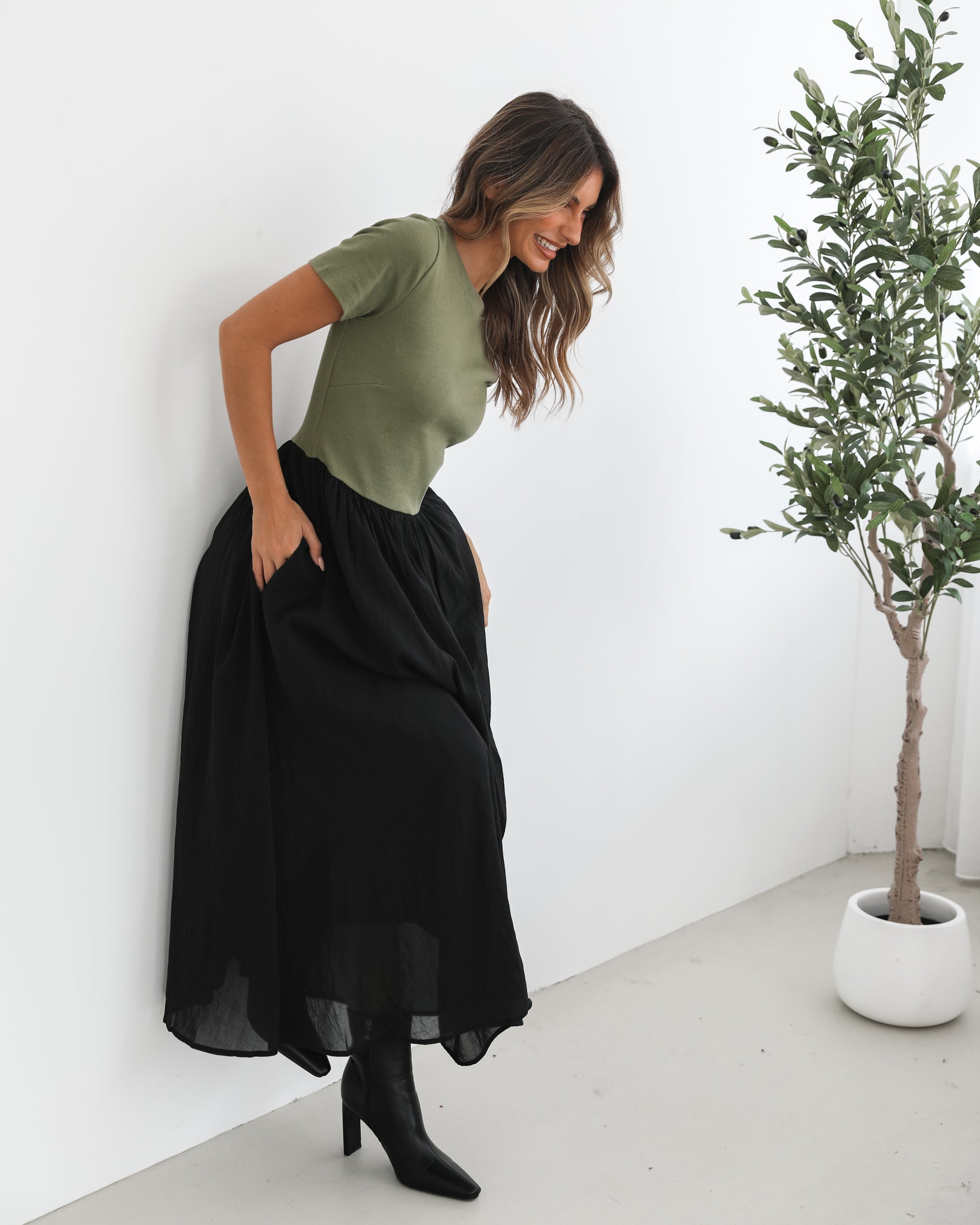 woman wearing a short sleeve round neck dress with green top and black flowy skirt paired with black boots in a white background studio