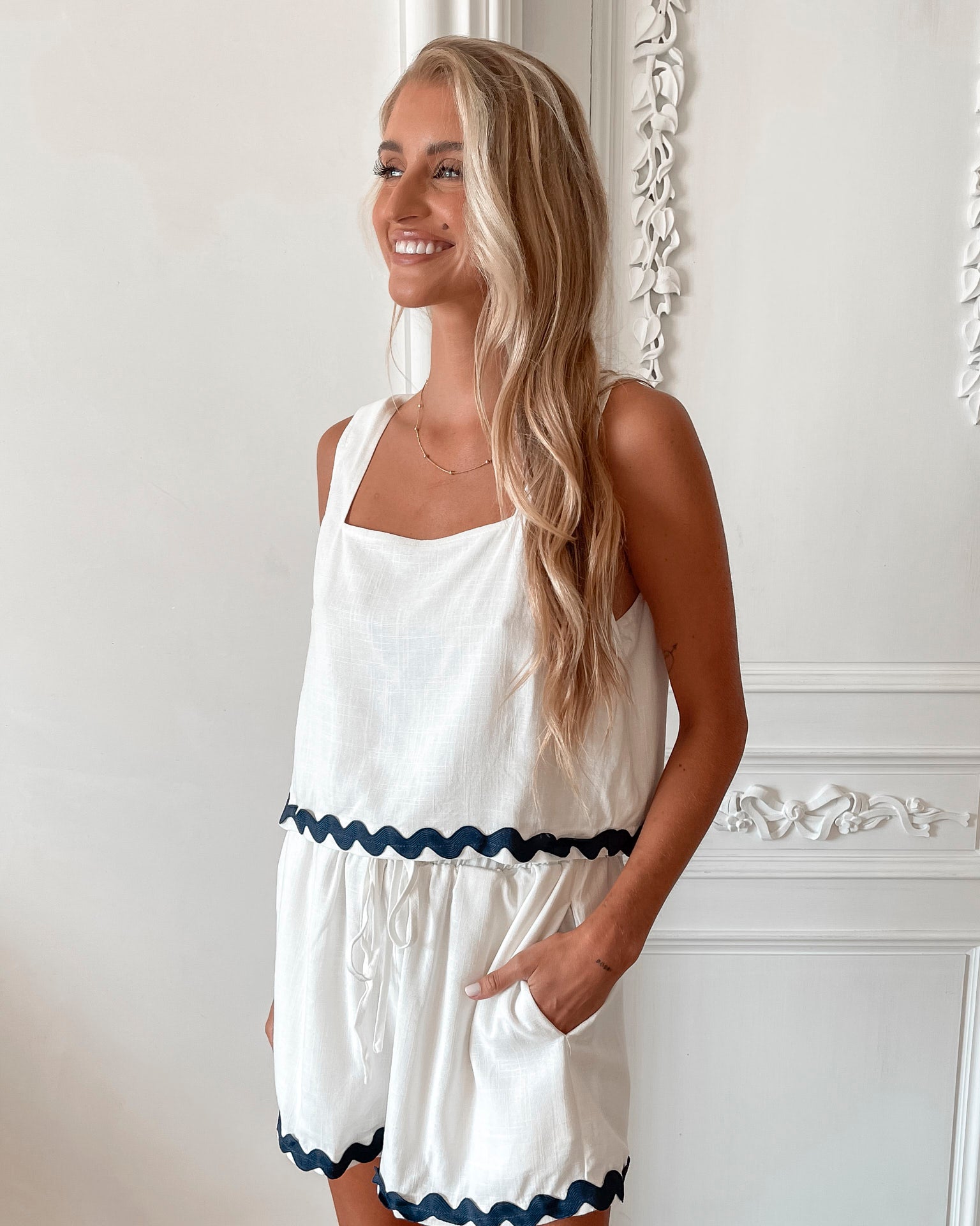 A woman is standing, wearing white high-waisted shorts with a string detail and side pockets, paired with a sleeveless top.