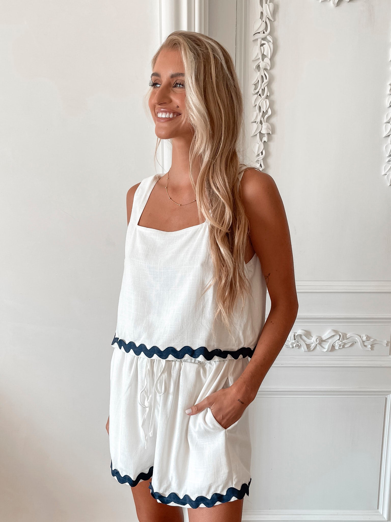 A woman is standing, wearing white high-waisted shorts with a string detail and side pockets, paired with a sleeveless top.
