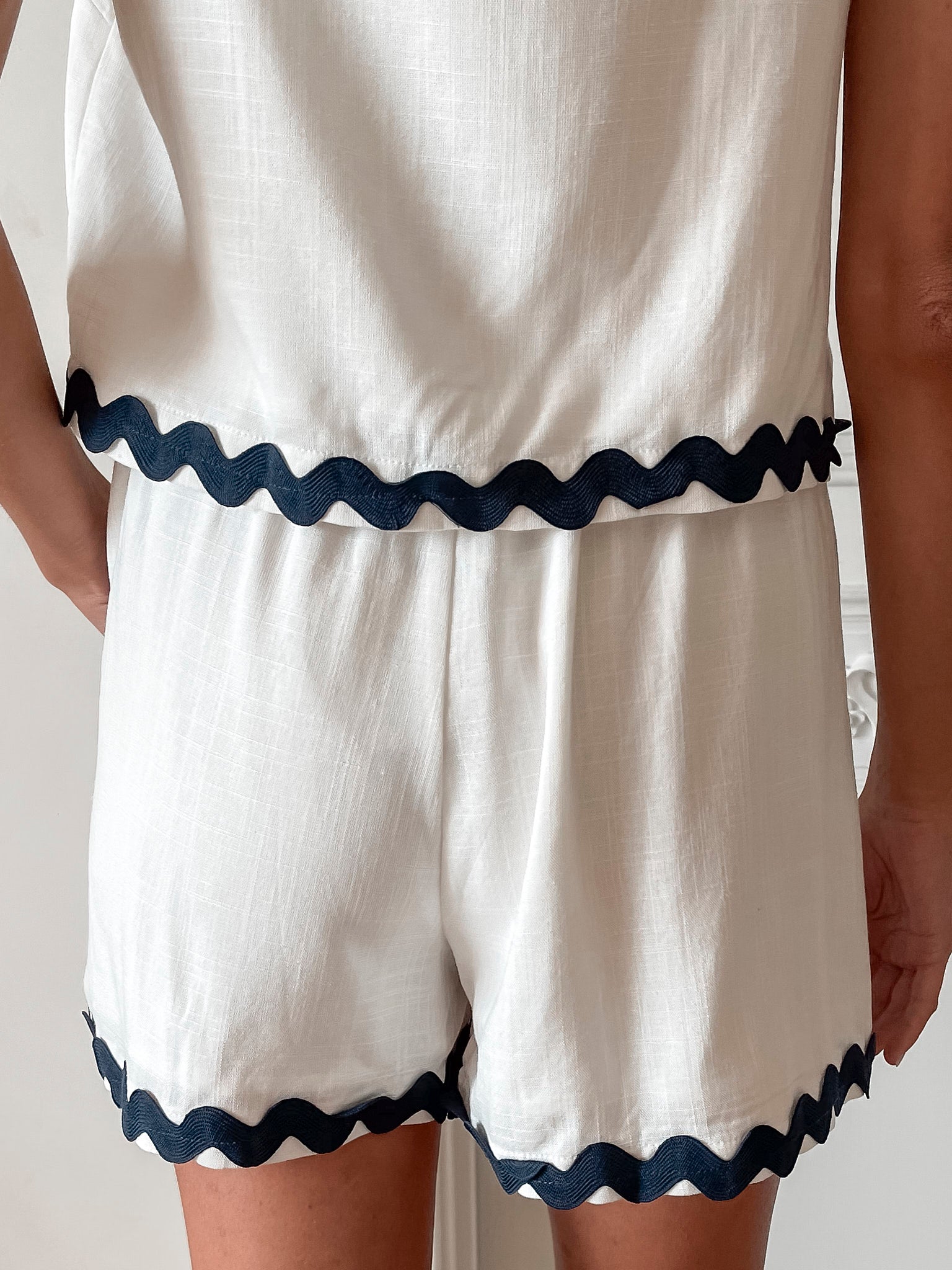 A woman is standing, wearing white high-waisted shorts with a string detail and side pockets, paired with a sleeveless top.