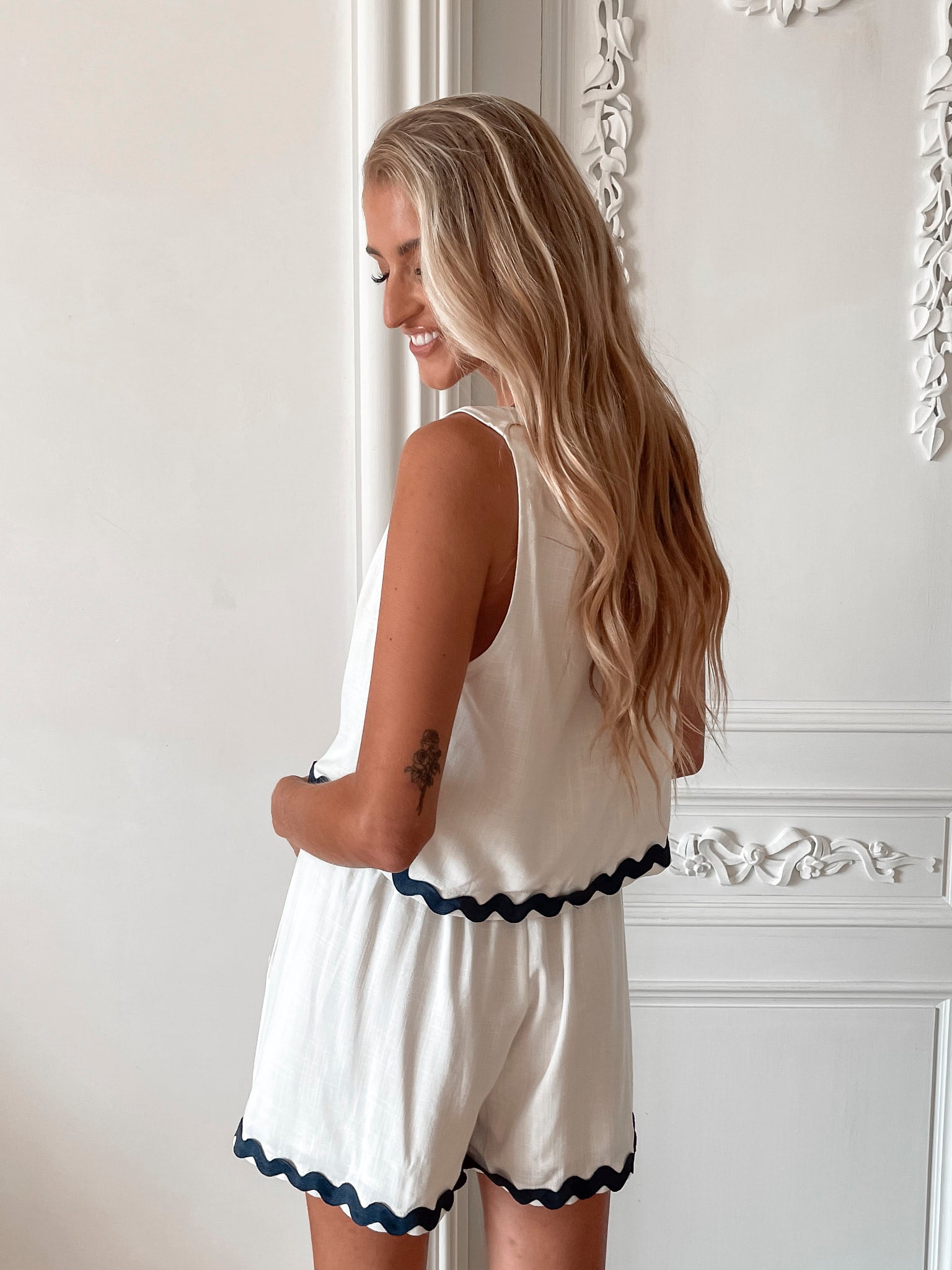 A woman is standing, wearing white high-waisted shorts with a string detail and side pockets, paired with a sleeveless top.