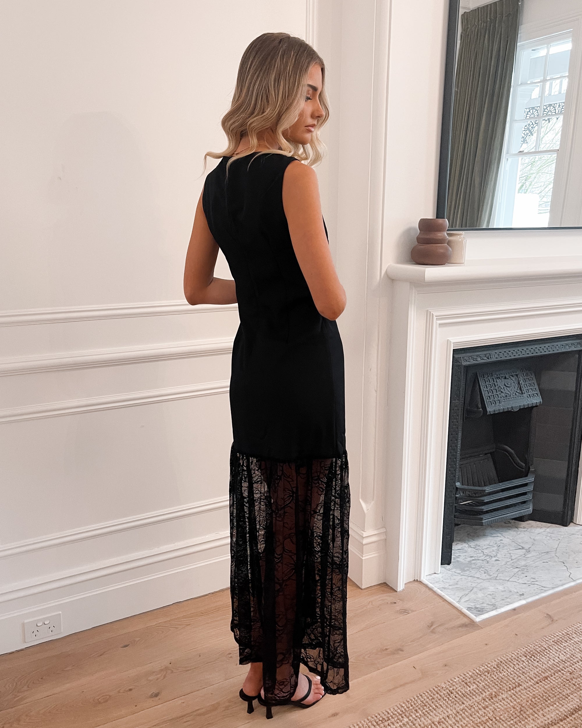 Chic woman in a black sleeveless dress with intricate lace overlay, perfect for black-tie events, weddings, evening galas, formal balls, and other elegant occasions, standing in a stylish room with wooden flooring and a fireplace.