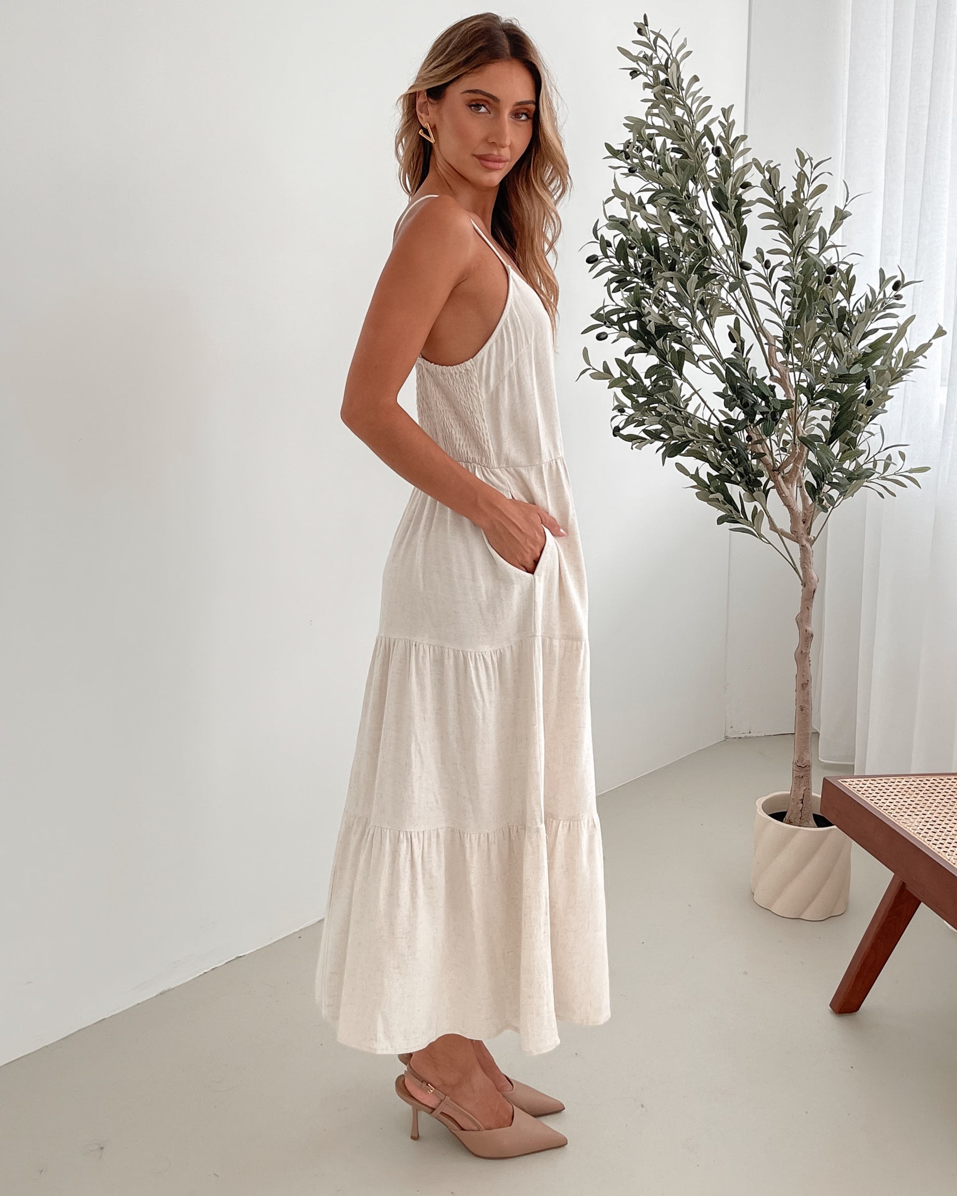 A woman wearing a beige, midi-length Sonya dress with a V neckline, adjustable spaghetti straps, a shirred back, and a tiered skirt. The dress is sleeveless and the photo shows the dress in a relaxed fit style.