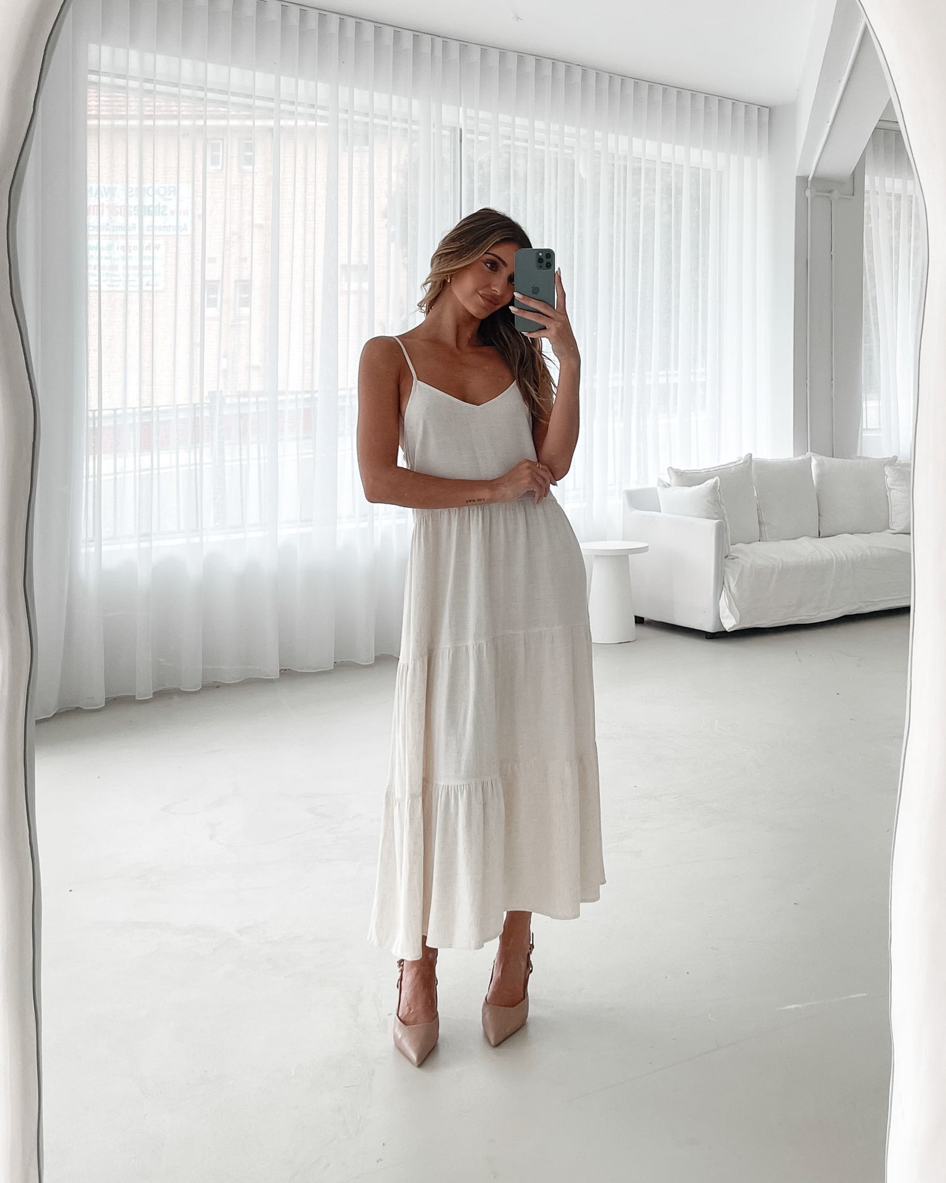 A woman wearing a beige, midi-length Sonya dress with a V neckline, adjustable spaghetti straps, a shirred back, and a tiered skirt. The dress is sleeveless and the photo shows the dress in a relaxed fit style.