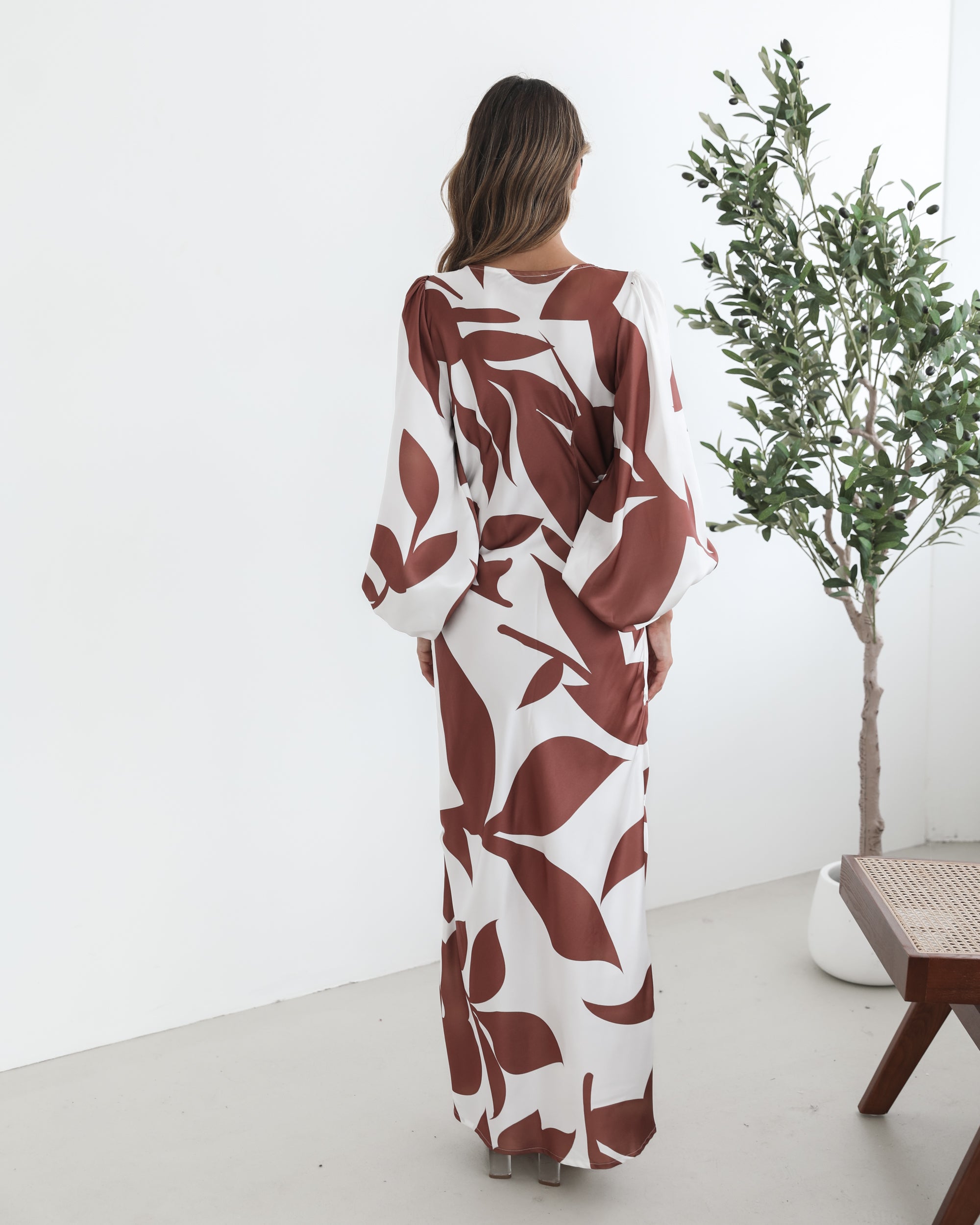 A model wearing a white and camel colored maxi dress with a wide v-neckline and long bishop sleeves, in a fit-to-flare silhouette with a maxi length hem.