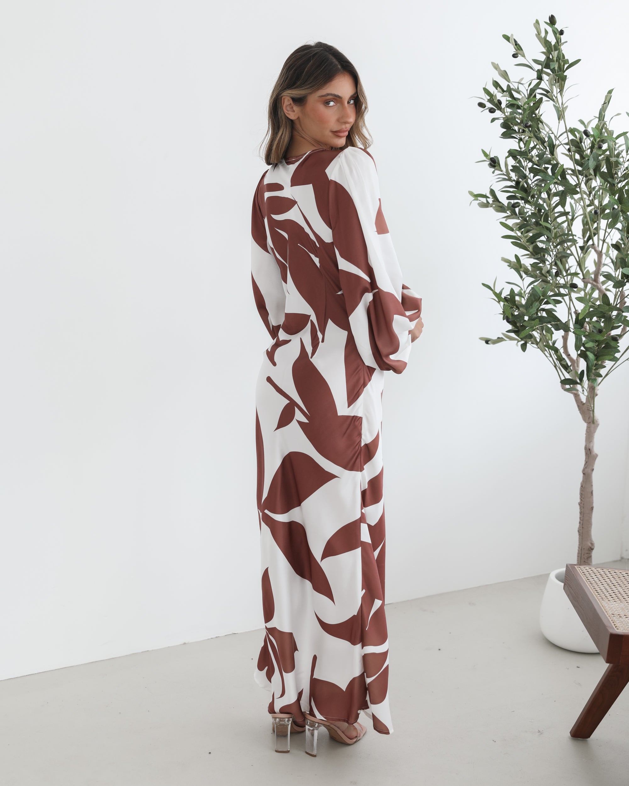 A model wearing a white and camel colored maxi dress with a wide v-neckline and long bishop sleeves, in a fit-to-flare silhouette with a maxi length hem.