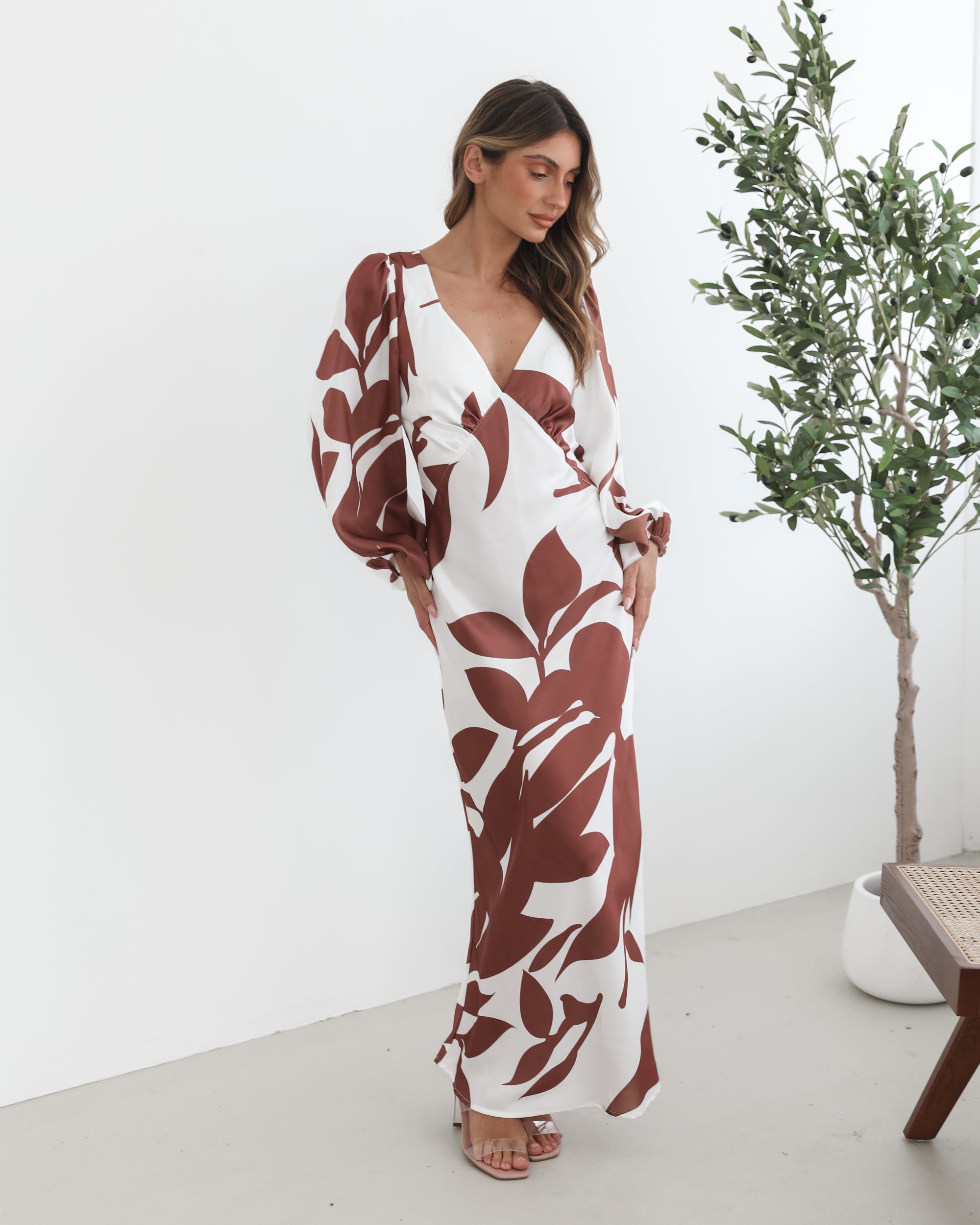 A model wearing a white and camel colored maxi dress with a wide v-neckline and long bishop sleeves, in a fit-to-flare silhouette with a maxi length hem.