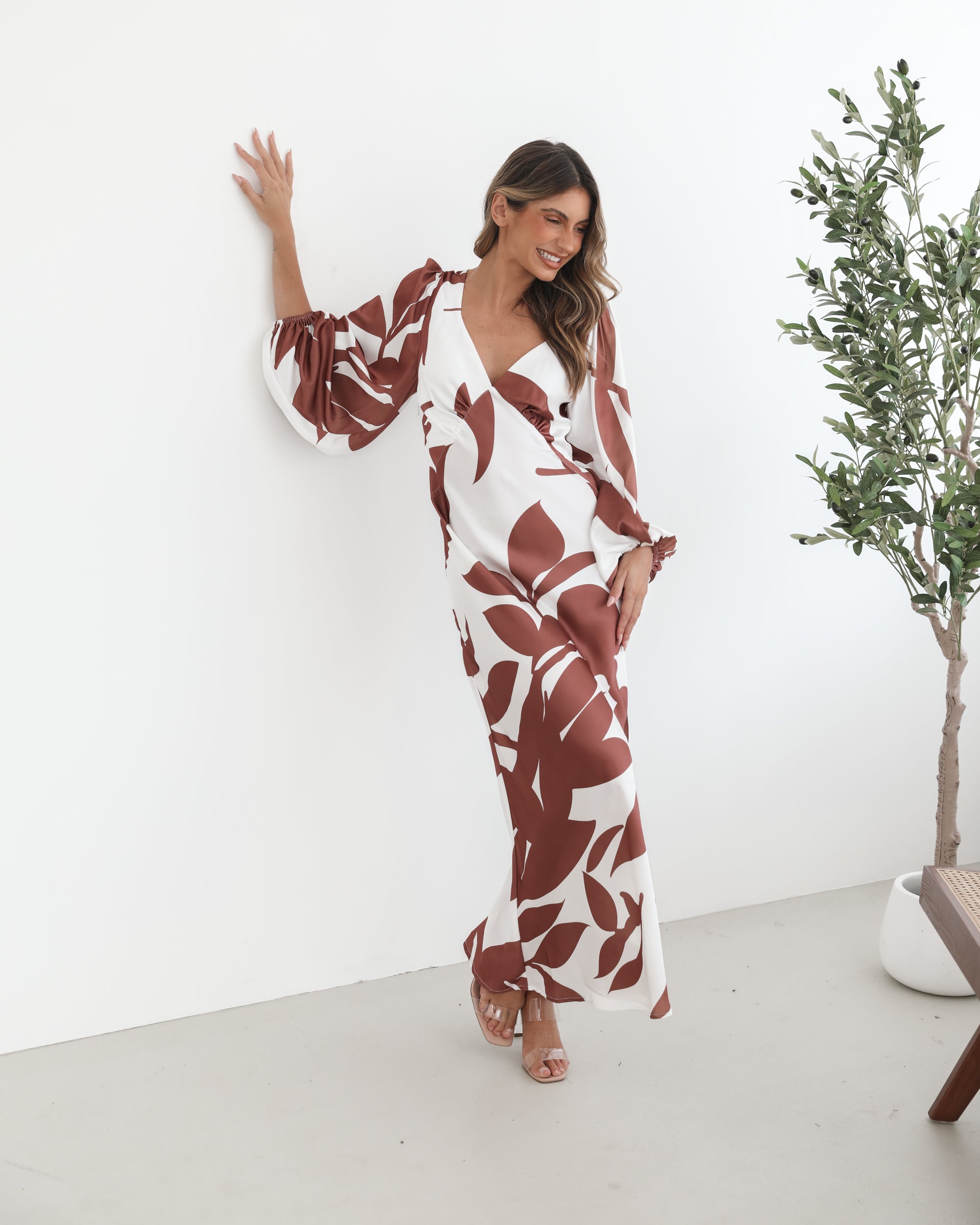 A model wearing a white and camel colored maxi dress with a wide v-neckline and long bishop sleeves, in a fit-to-flare silhouette with a maxi length hem.
