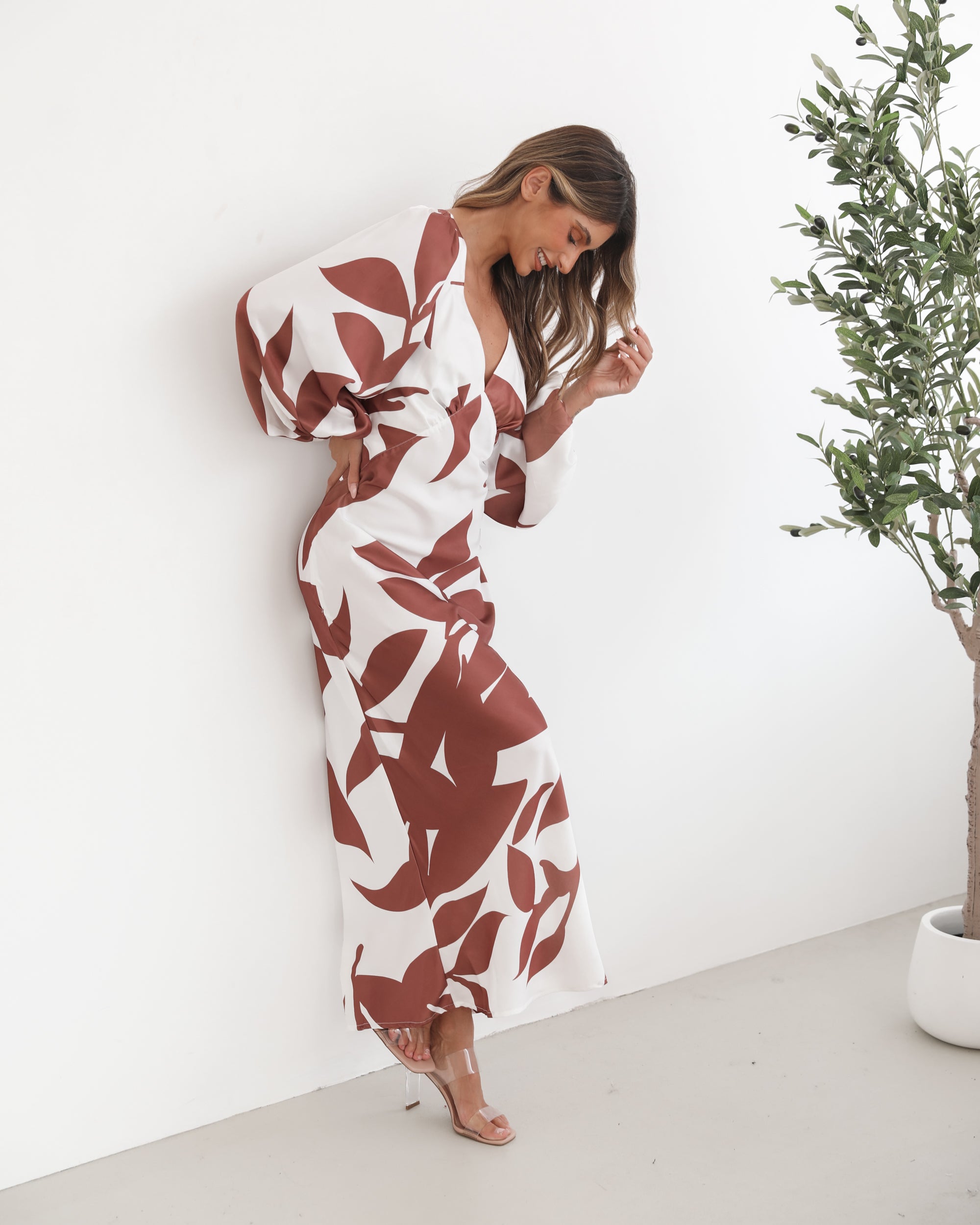 A model wearing a white and camel colored maxi dress with a wide v-neckline and long bishop sleeves, in a fit-to-flare silhouette with a maxi length hem.