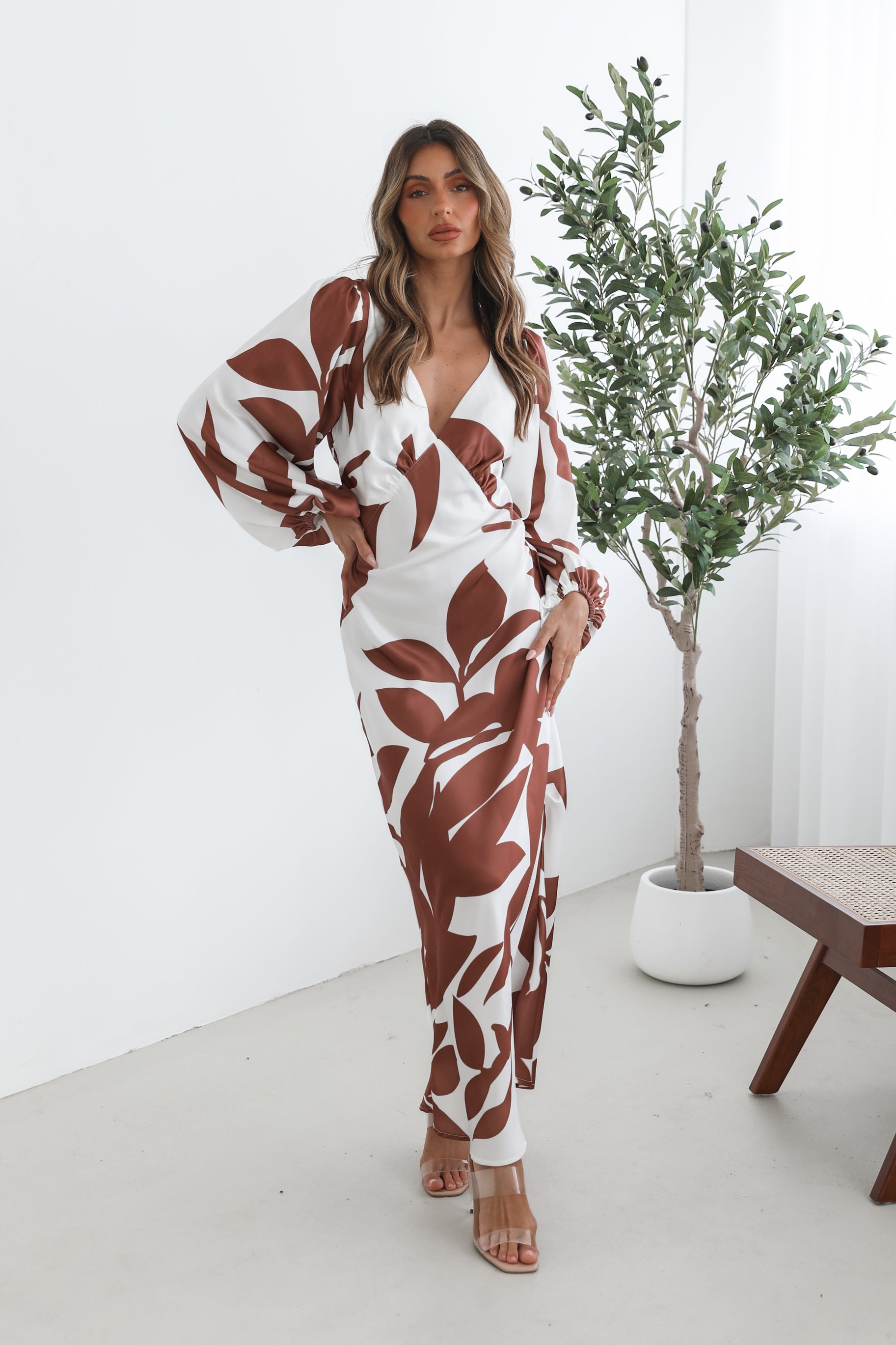 A model wearing a white and camel colored maxi dress with a wide v-neckline and long bishop sleeves, in a fit-to-flare silhouette with a maxi length hem.