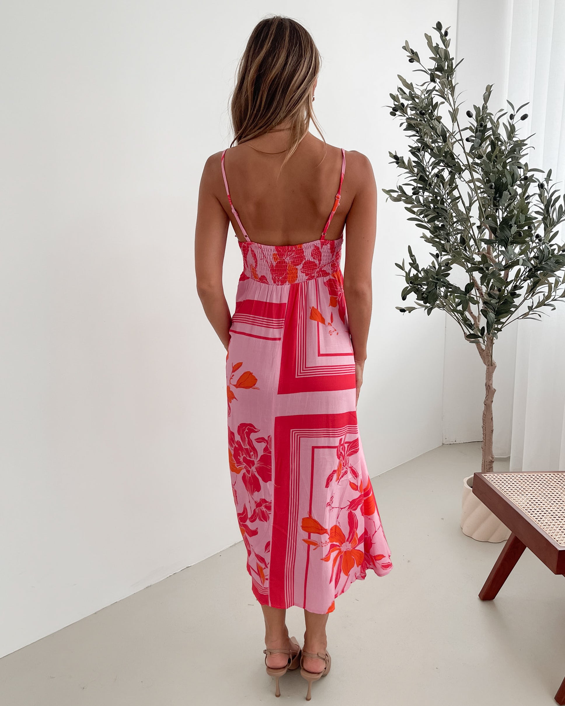 A woman wearing a pink floral dress with spaghetti straps and a square neckline. The dress has a flowy a-line skirt and she is standing in a room with a plant in the background.
