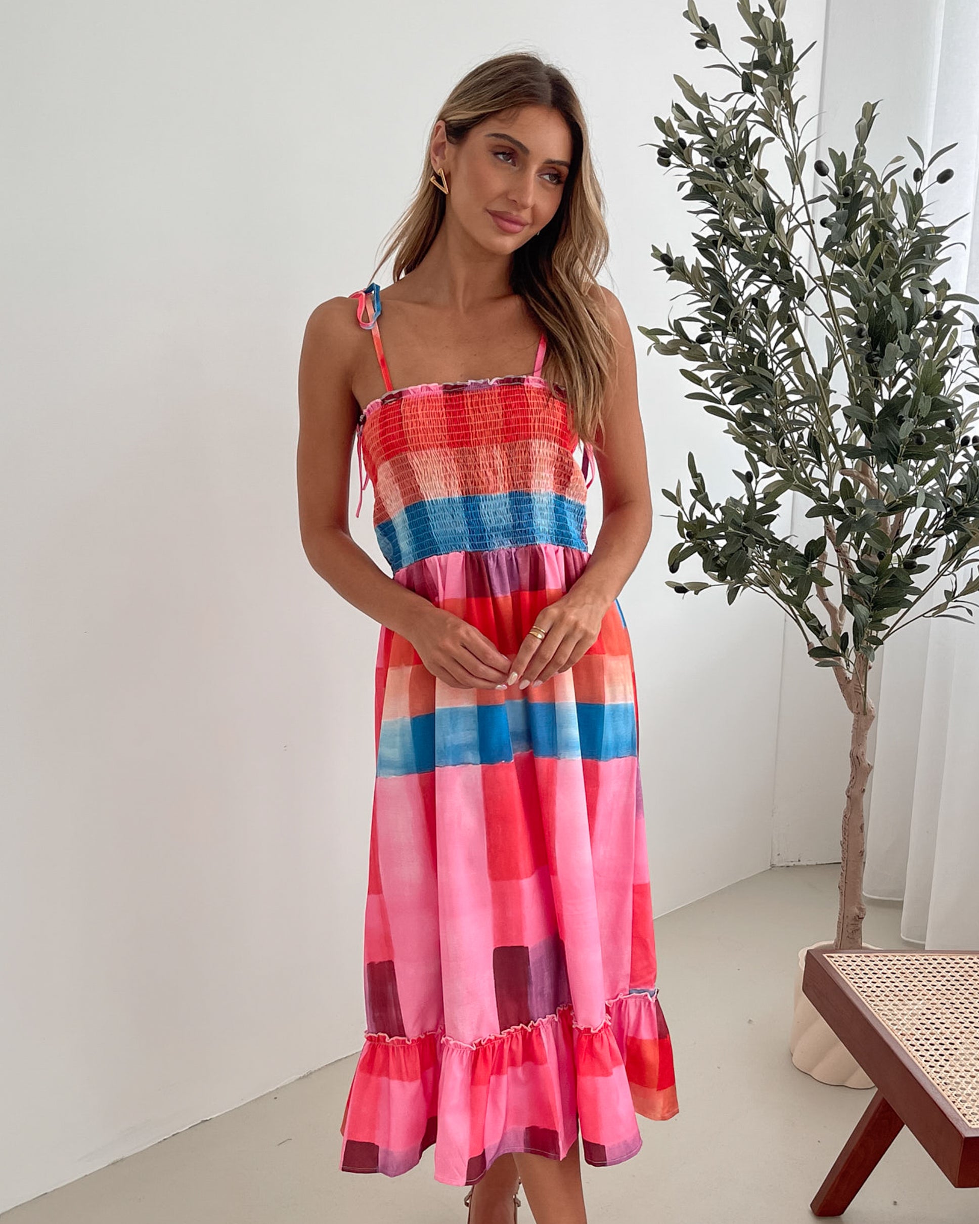 A woman standing in a room with a white wall and a small wooden chair, wearing a multicolored sleeveless dress with a square neckline and spaghetti straps. The dress features a smocked bodice and a flowy skirt.
