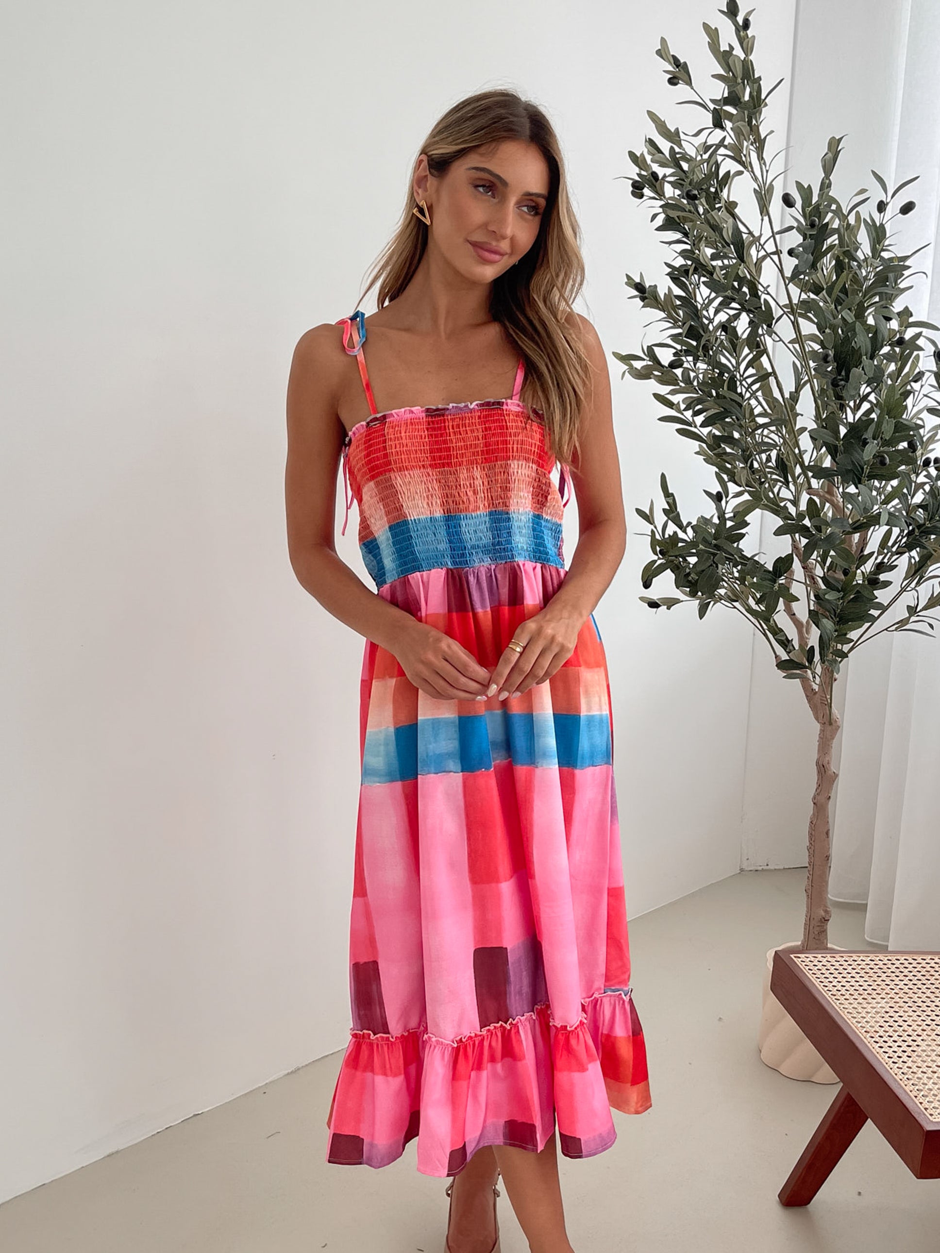 A woman standing in a room with a white wall and a small wooden chair, wearing a multicolored sleeveless dress with a square neckline and spaghetti straps. The dress features a smocked bodice and a flowy skirt.