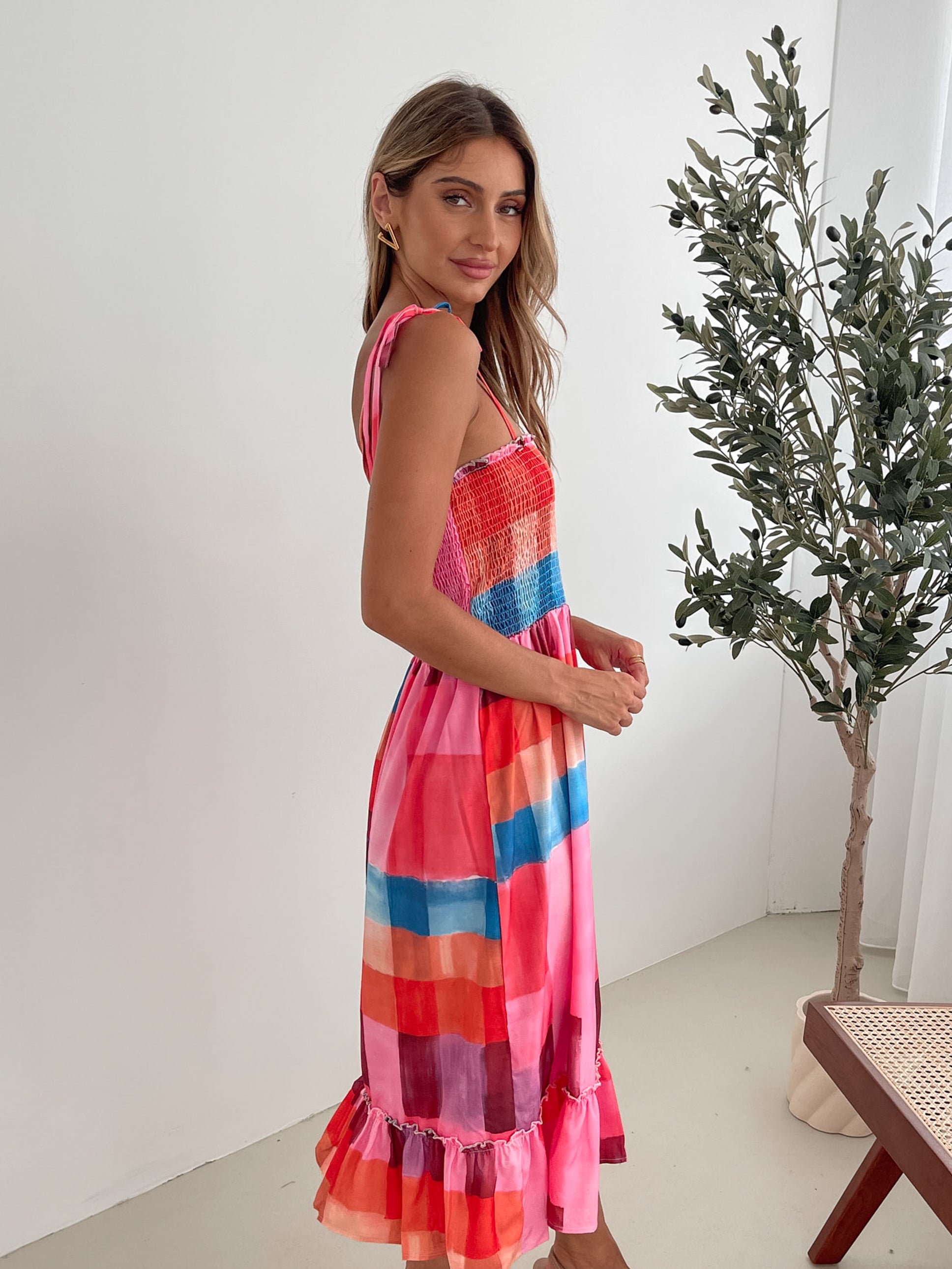 A woman standing in a room with a white wall and a small wooden chair, wearing a multicolored sleeveless dress with a square neckline and spaghetti straps. The dress features a smocked bodice and a flowy skirt.