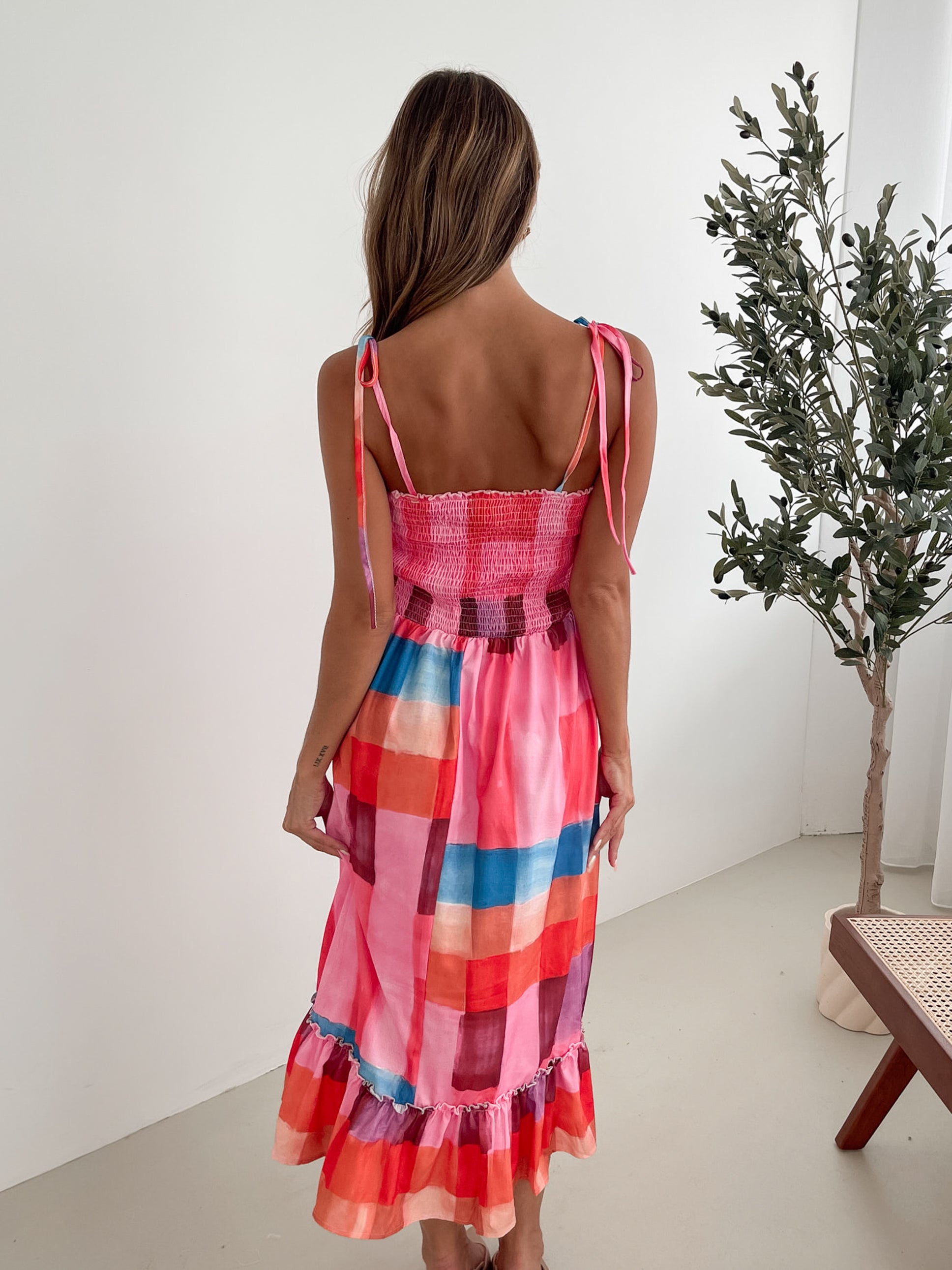 A woman standing in a room with a white wall and a small wooden chair, wearing a multicolored sleeveless dress with a square neckline and spaghetti straps. The dress features a smocked bodice and a flowy skirt.